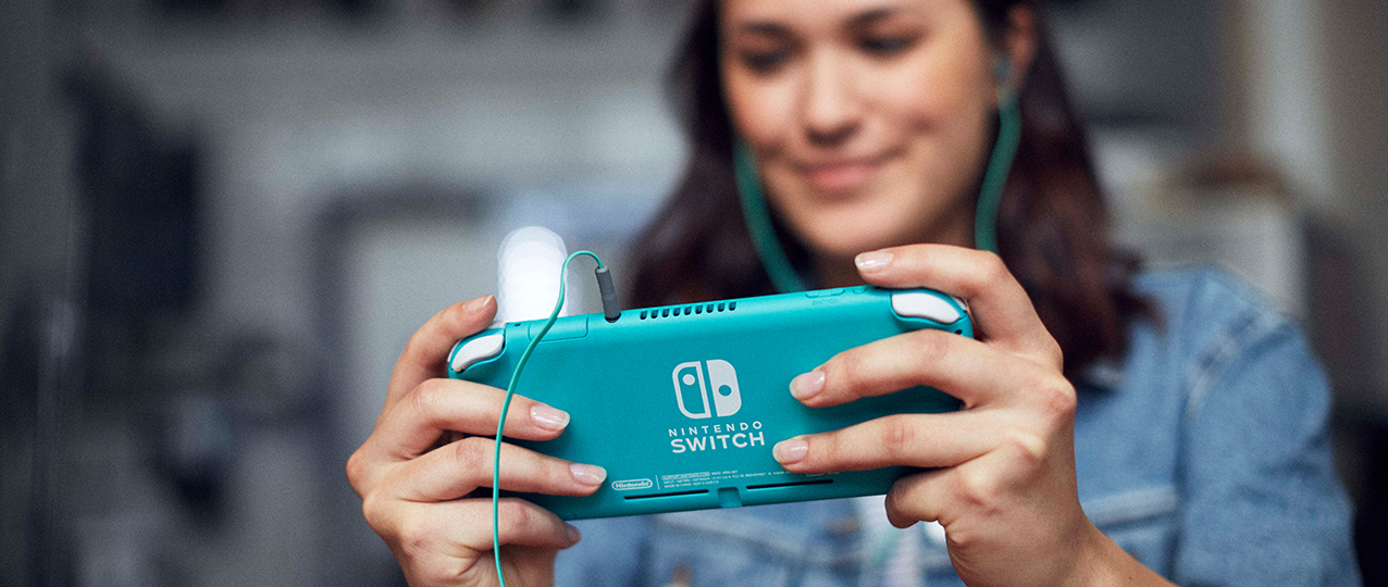 Eine Frau hält eine türkisfarbene Nintendo Switch Lite mit Kopfhörern. Fokus auf Konsole und Hände. Der Hintergrund ist unscharf.