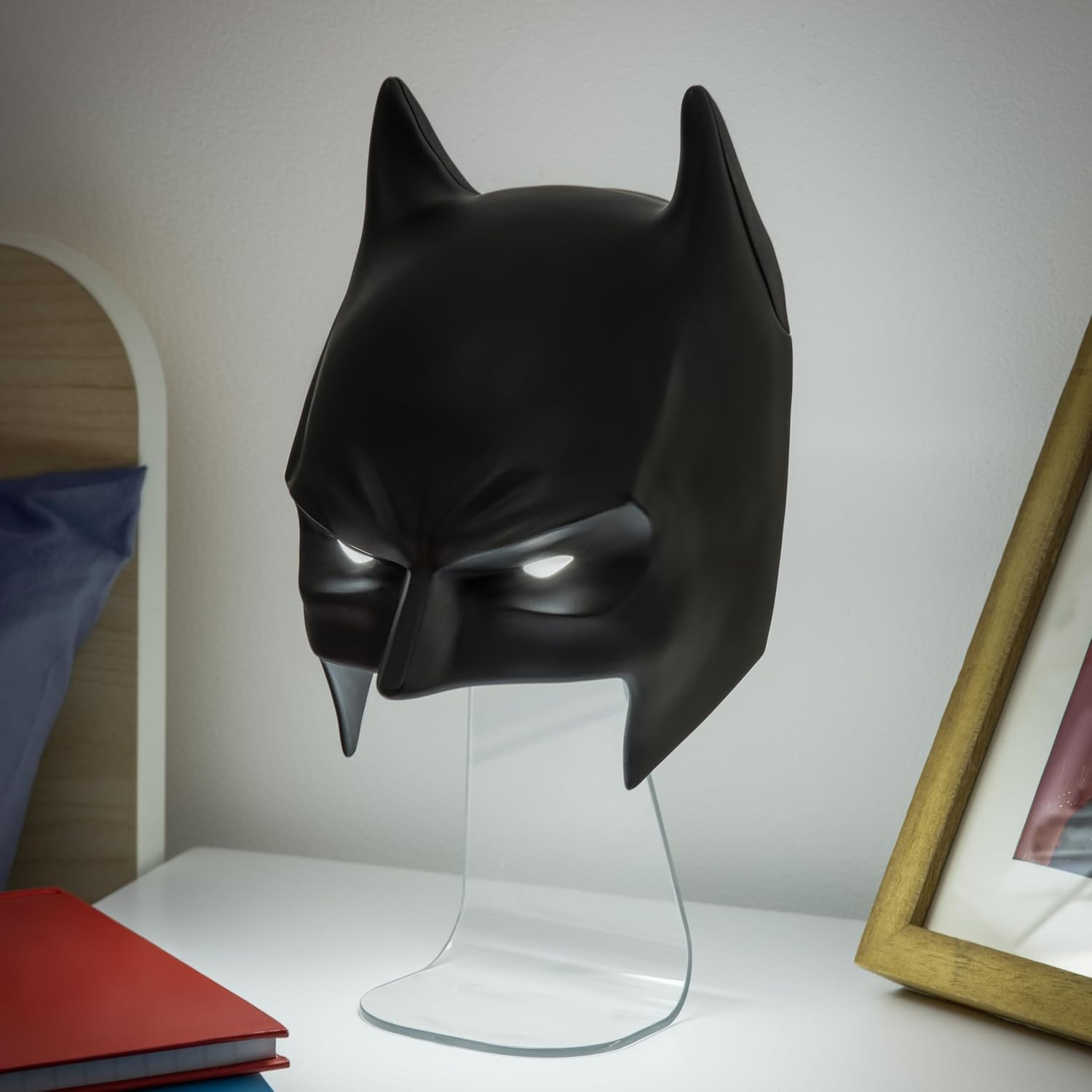 Egy Batman maszk lámpa egy fehér asztalon. A maszk fekete, világító szemekkel. Közelben egy képkeret és könyvek láthatók.