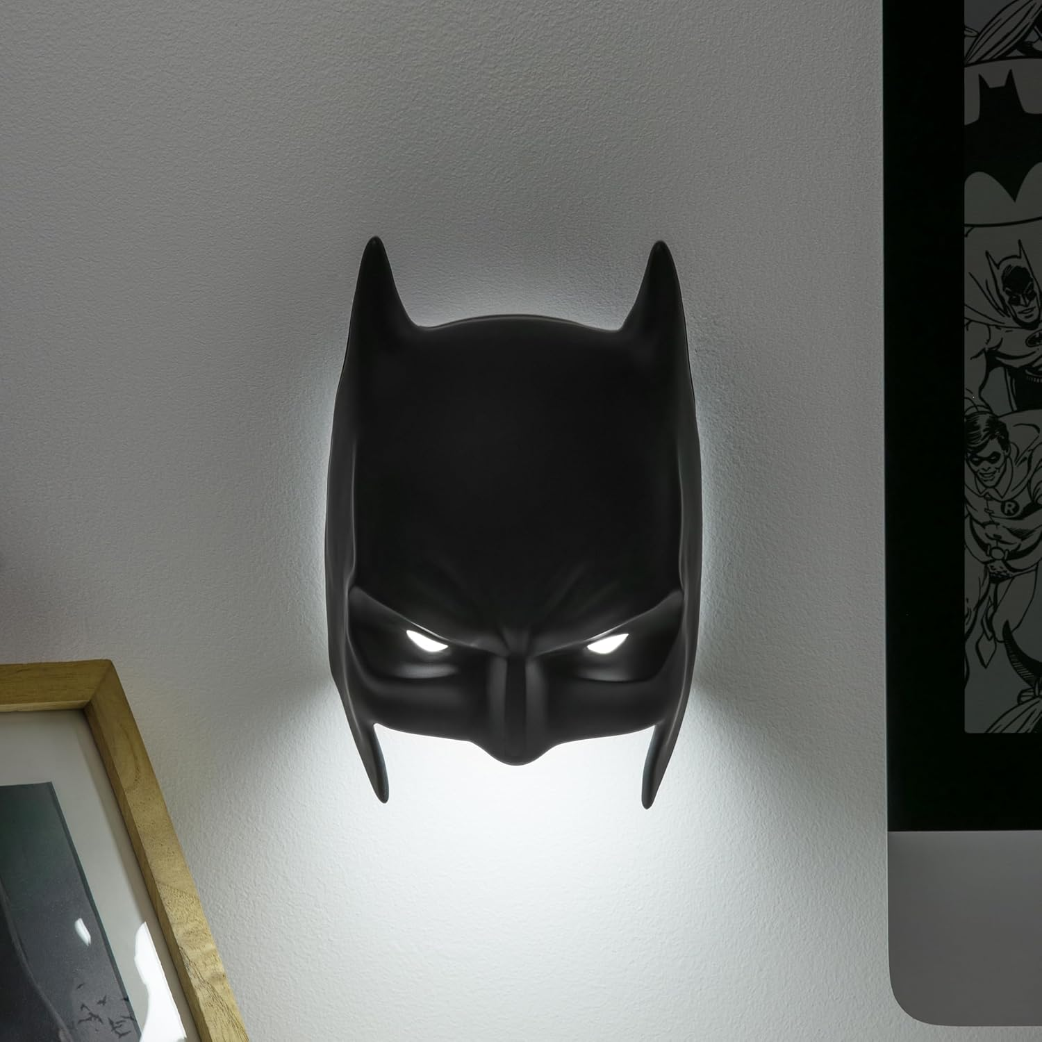 Fekete Batman maszk alakú fali lámpa fehér falon. Képregény a jobb oldalon. Egy képkeret bal oldalon.