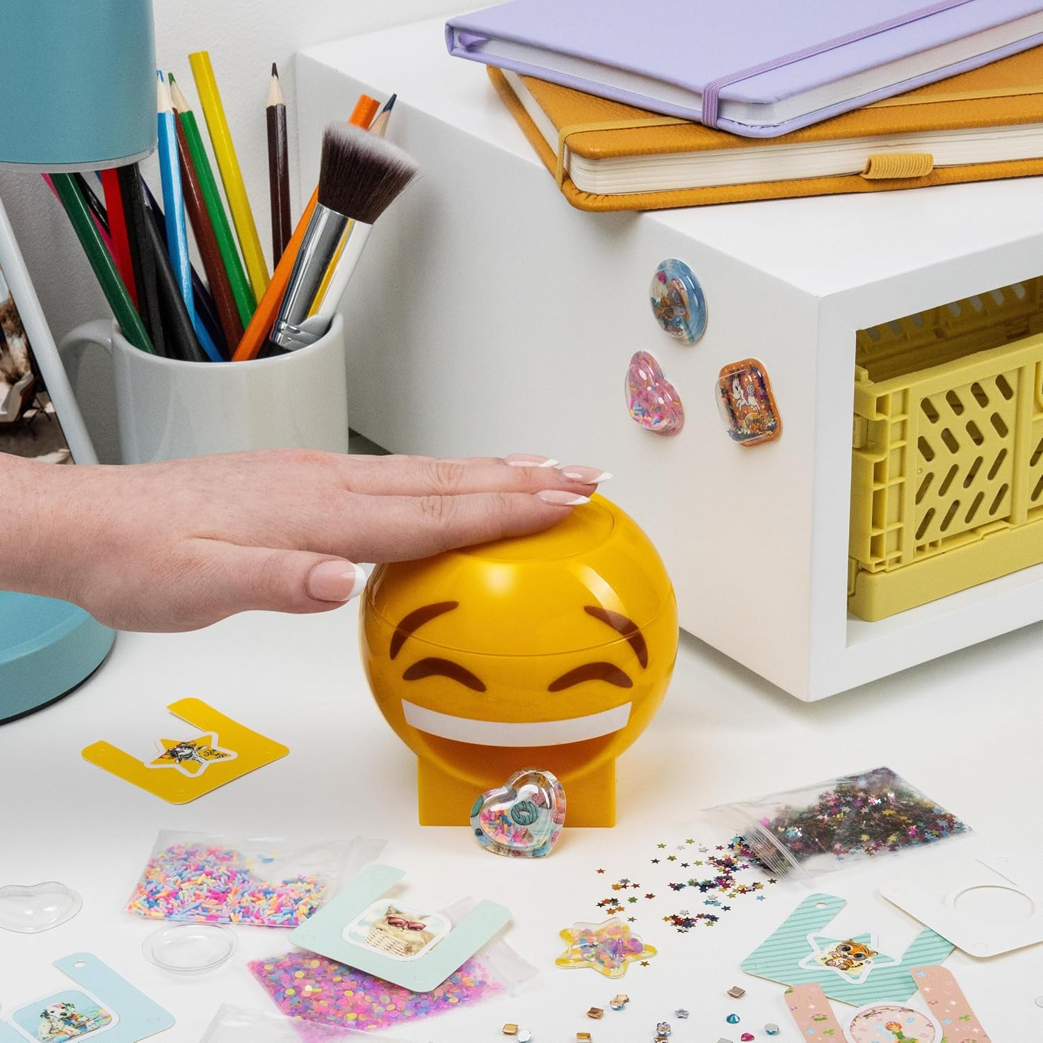 Egy kéz megérint egy sárga, emoji alakú tartályt. Színes ceruzák és egy ecset van egy bögrében. Egyéb tárgyak: jegyzetfüzetek és kézműves kellékek.