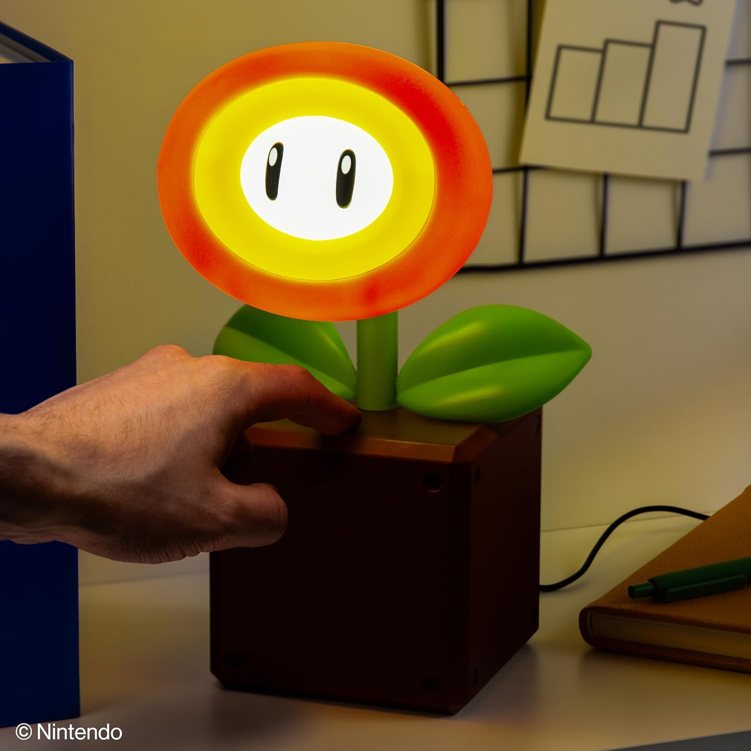 Egy kéz megérinti a Mario Tűzviráglámpát. A lámpa narancssárga, sárga és zöld. A háttérben egy kék tárgy és egy jegyzetfüzet látható.