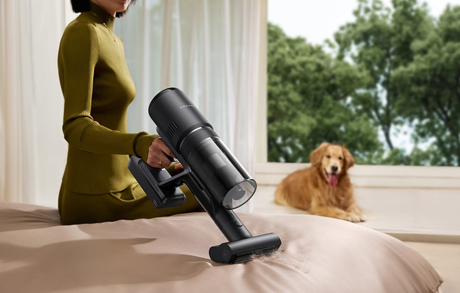 Egy nő fekete porszívót használ az ágyon. Egy golden retriever kutya ül a háttérben az ablaknál.