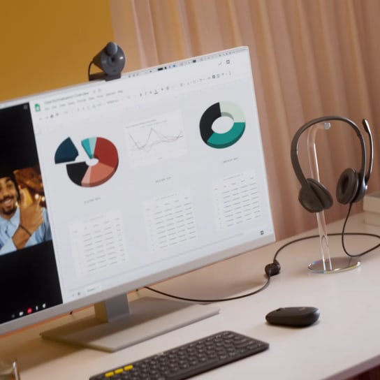 A számítógép monitor grafikonokat jelenít meg. Egy webkamera van rajta. Fejhallgató, egér és billentyűzet van az asztalon.