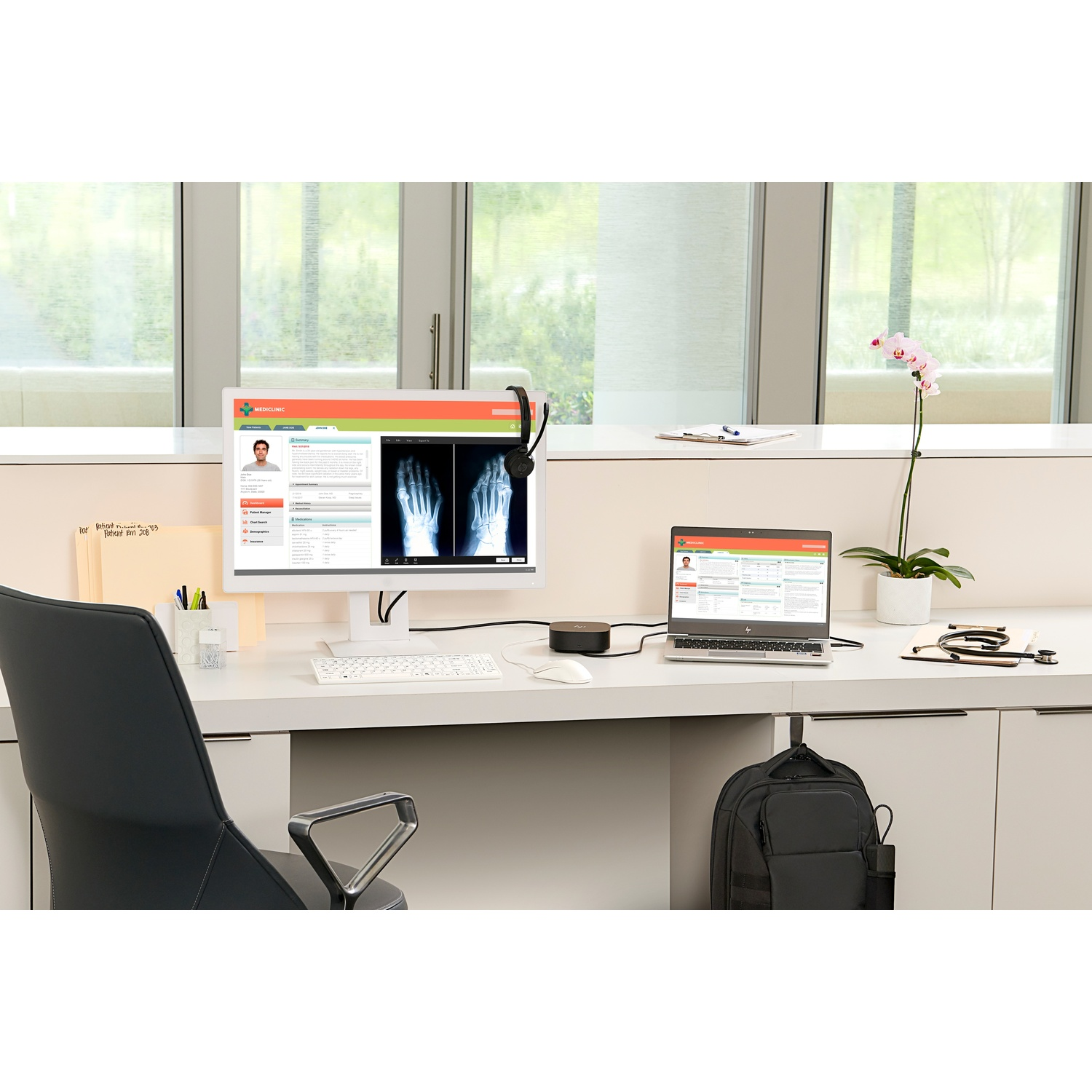 A medical office desk with a computer, laptop, and a headset. An orchid and a black bag are also on the desk.