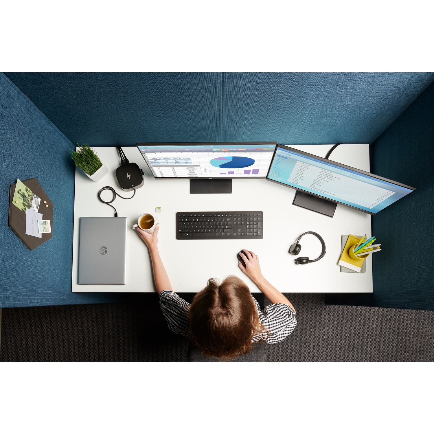 A person sits at a desk with dual monitors, laptop, keyboard, and mouse. They hold a cup. The desk is surrounded by blue walls.