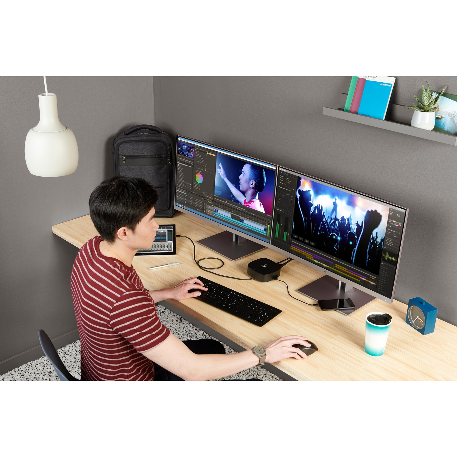 Man at desk with dual monitors editing video. Includes keyboard, mouse, phone, coffee cup, clock, backpack, and a hanging lamp.