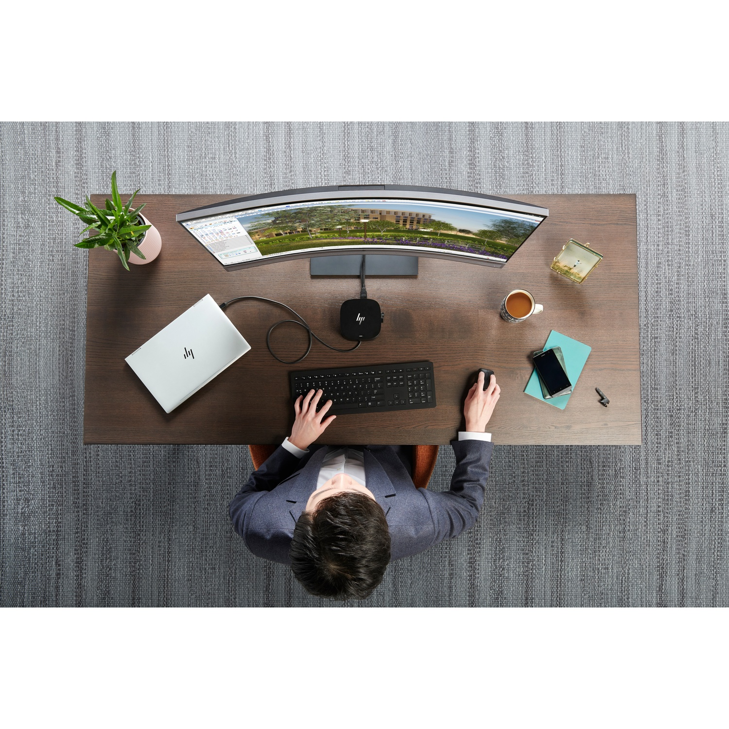 Overhead view of a person working at a desk with a curved monitor, laptop, keyboard, and other office items.