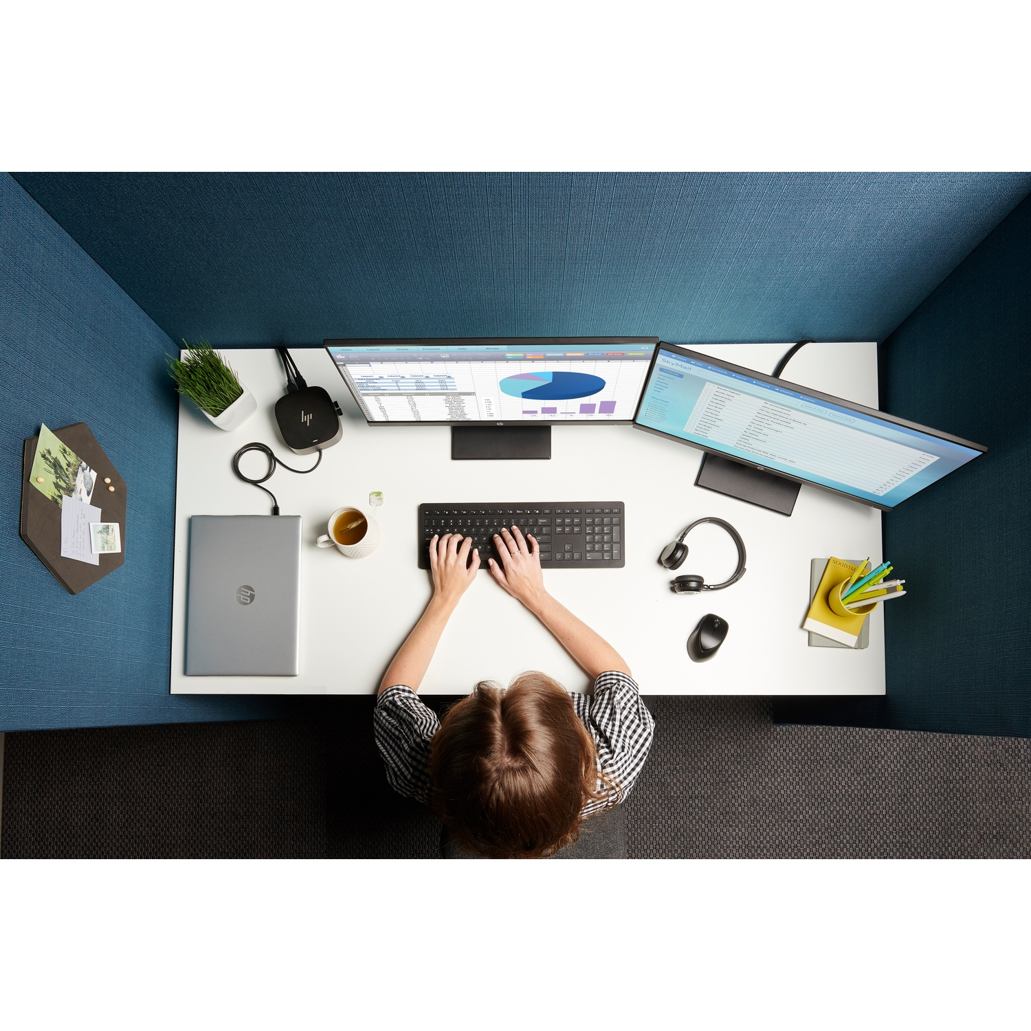 A person working at a desk with two monitors, keyboard, laptop, and other office supplies. The desk is in a cubicle setting.