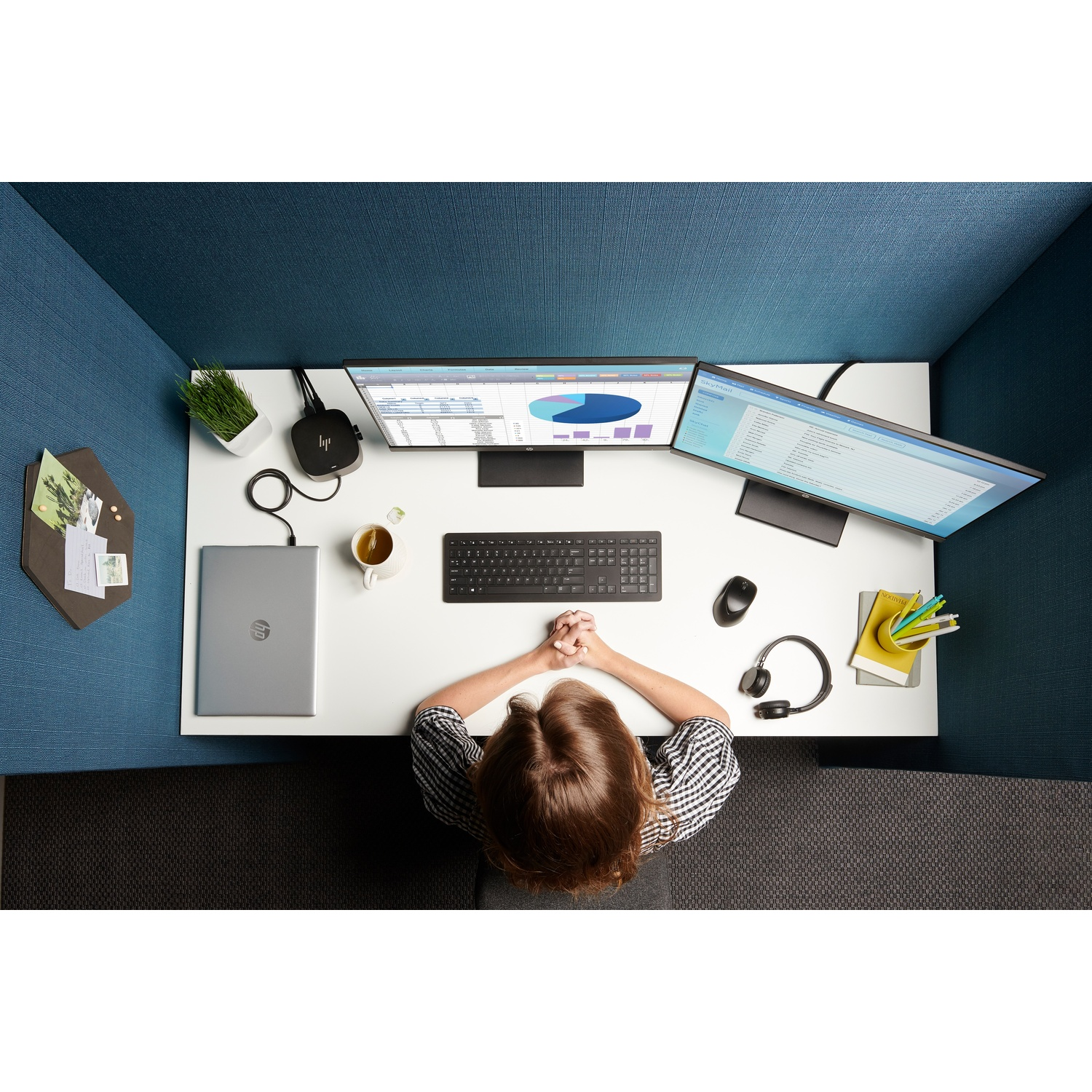 Overhead view of a workspace. Woman at desk with dual monitors, keyboard, mouse, headphones, and laptop. Blue textured wall.