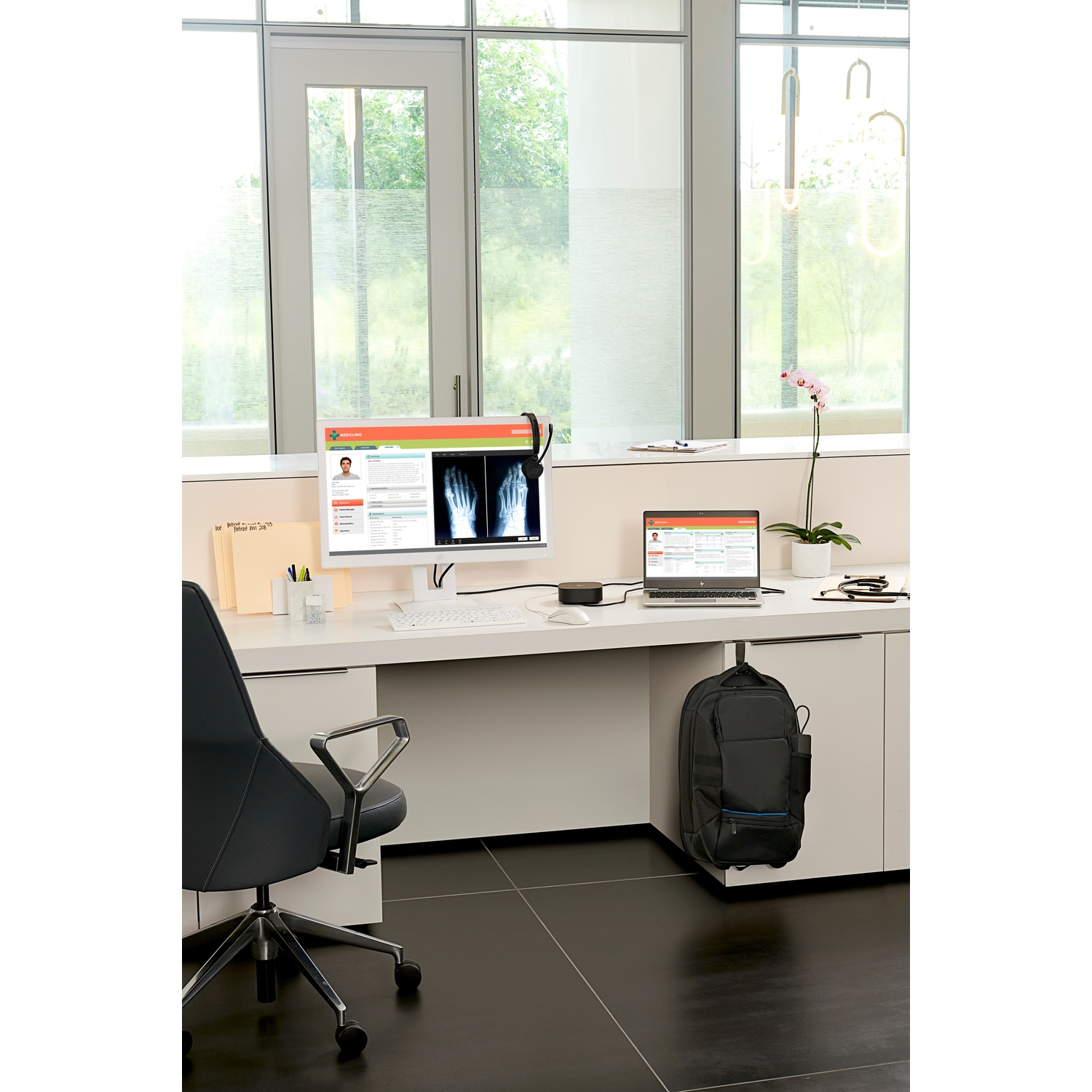 A doctor's office with a computer displaying X-rays. A laptop sits beside it. A black chair and backpack are in the scene.