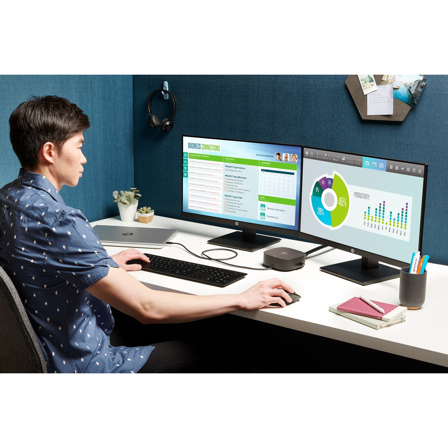 A man sits at a desk with two monitors, using a mouse and keyboard. The monitors display data and charts.