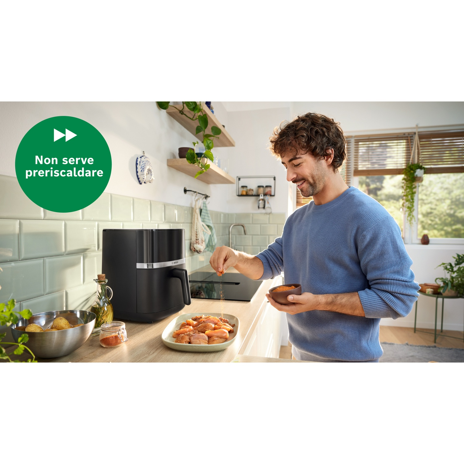 Un uomo con un maglione blu condisce il cibo su un piatto. Una friggitrice ad aria nera, verdure e utensili da cucina sono visibili.