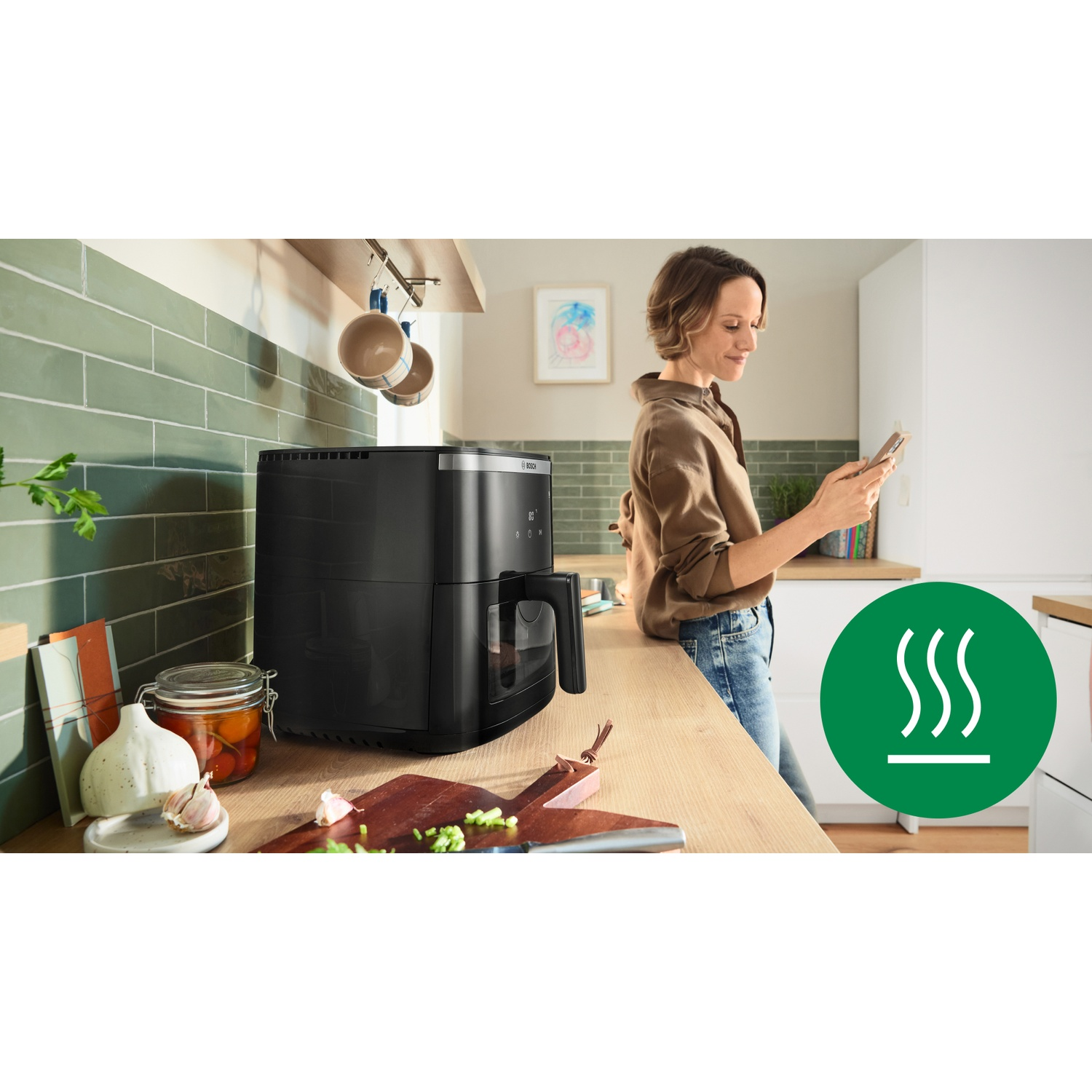 Una donna in cucina guarda il telefono. Una friggitrice ad aria nera è su un bancone di legno. Piastrelle verdi e armadi bianchi sullo sfondo.