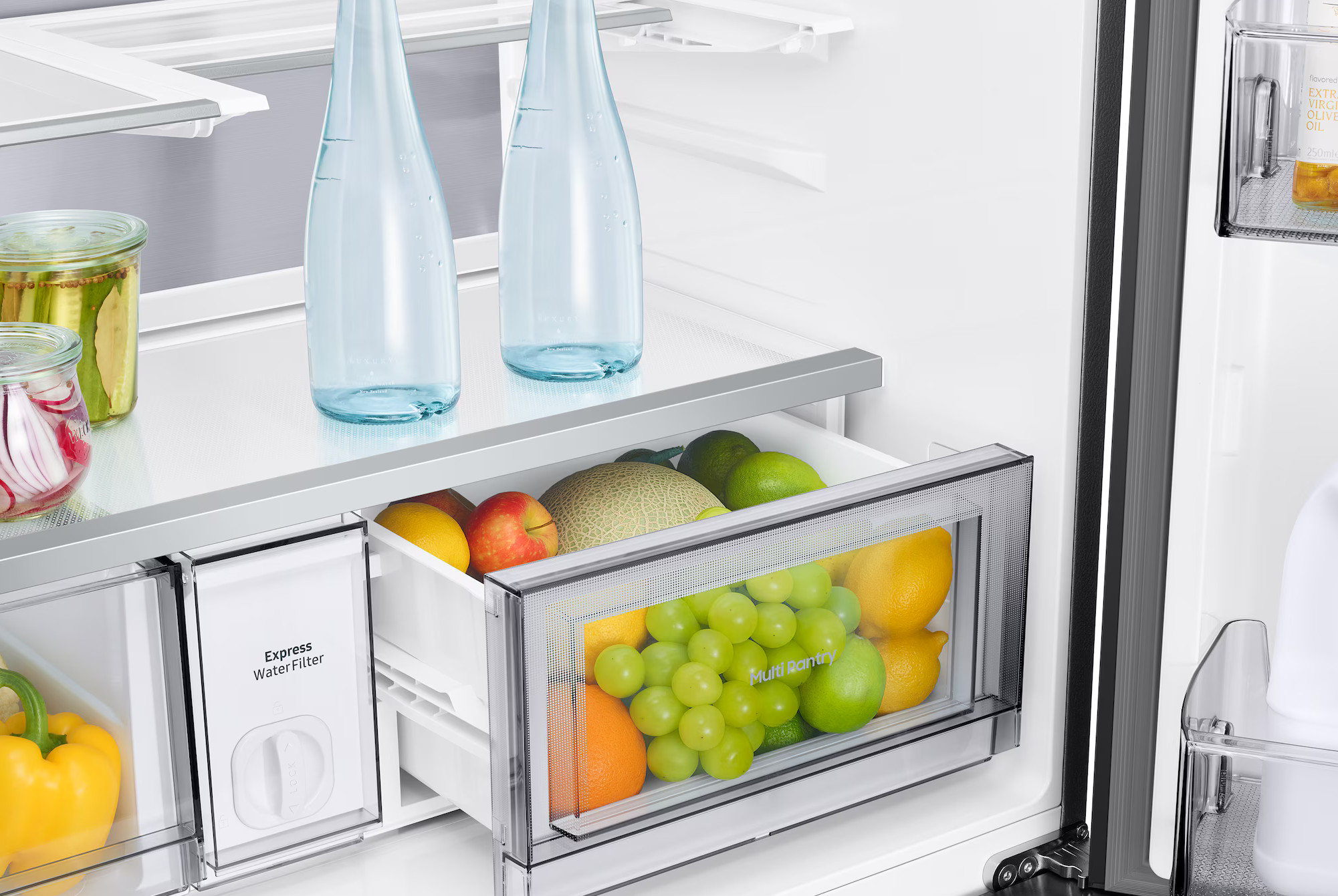 Wewnątrz lodówki: butelki z wodą, owoce takie jak winogrona, cytryny, jabłka i melon oraz słoiki z ogórkami i rzodkiewkami.