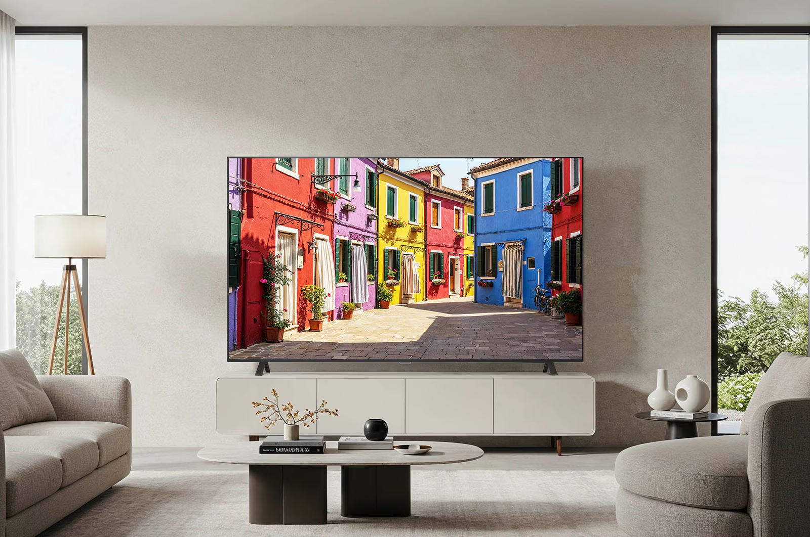 Ein Wohnzimmer mit einem Fernseher, der farbenfrohe Gebäude zeigt. Es gibt ein Sofa, eine Lampe und einen Couchtisch. Große Fenster zeigen eine grüne Aussicht.