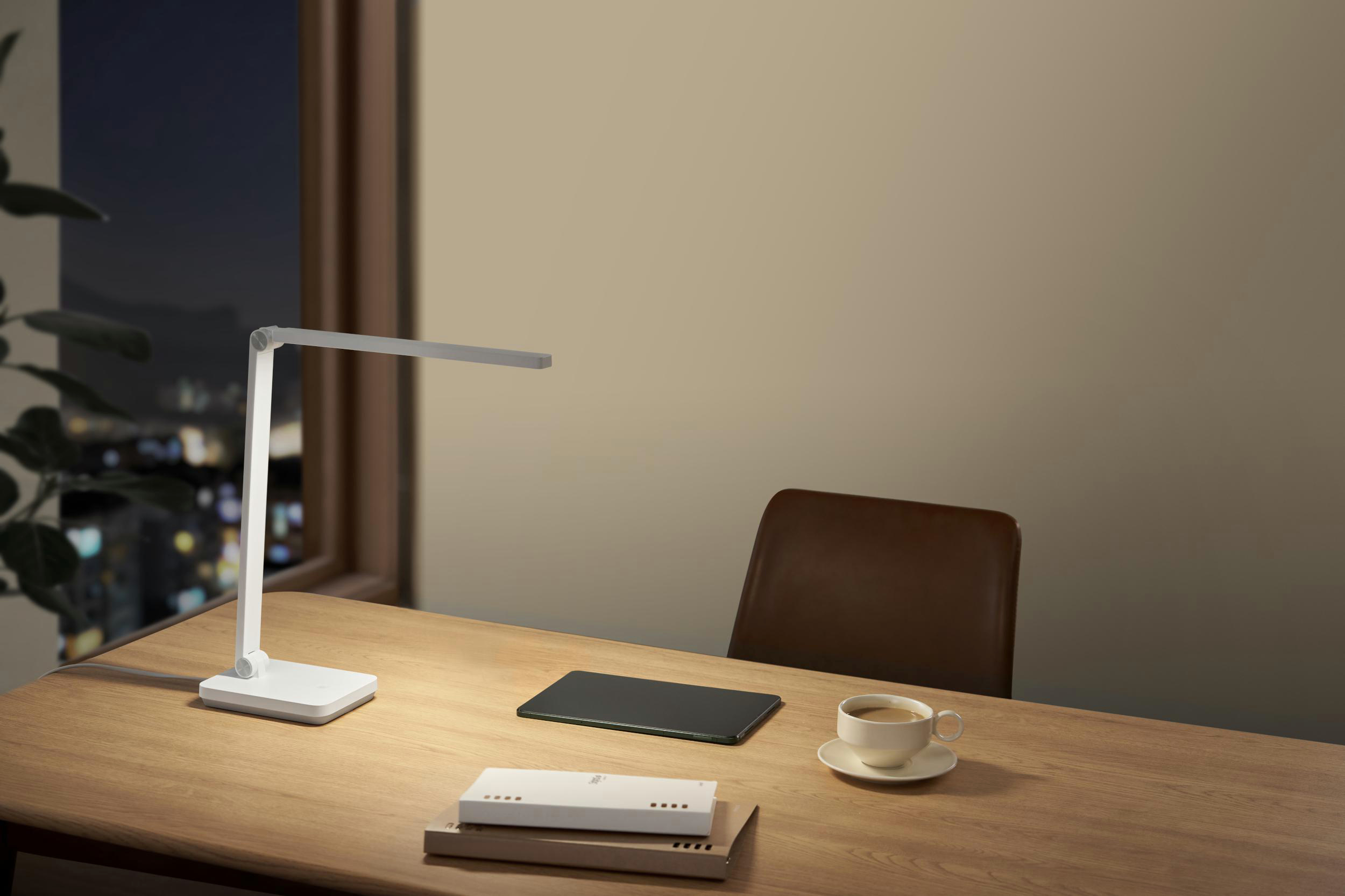 Ein Schreibtisch mit Lampe, Tablet, Kaffeetasse und Büchern. Ein Stuhl steht hinter dem Schreibtisch. Durchs Fenster sind Stadtlichter zu sehen.