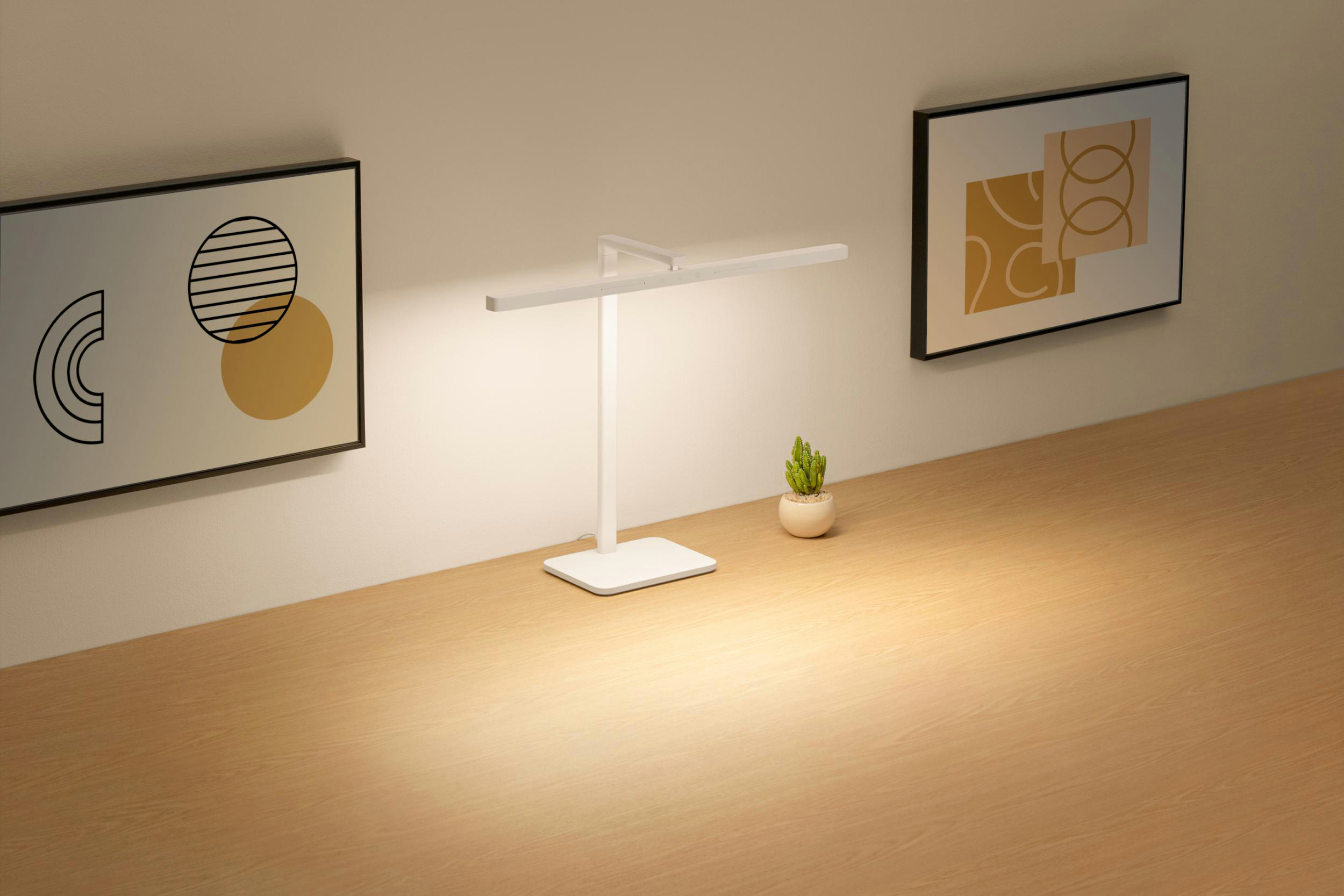 Eine weiße Schreibtischlampe, ein Kaktus und Kunstdrucke auf einem Holztisch. Beige Wände. Geometrische Kunst.
