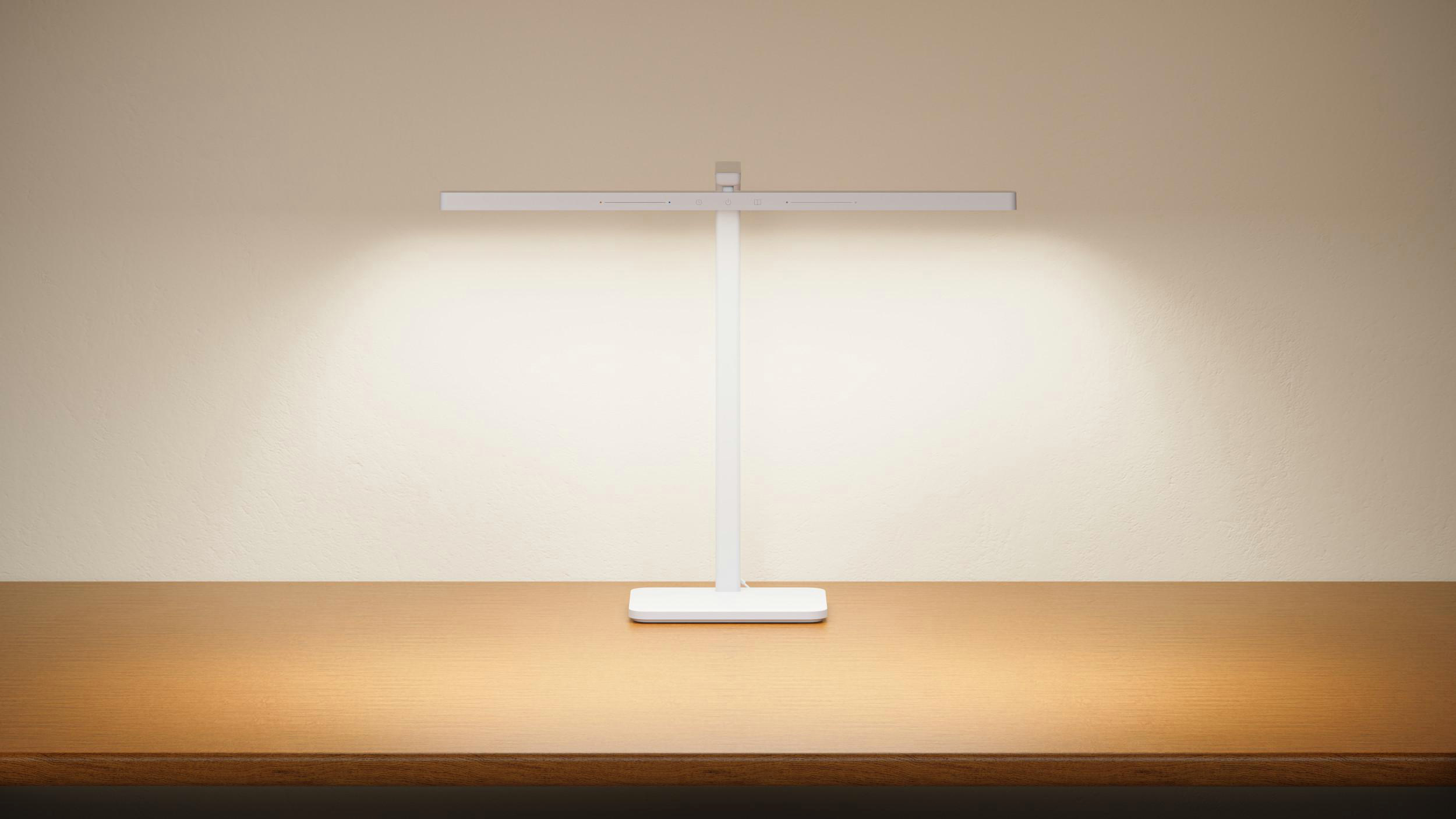 Eine weiße Schreibtischlampe steht auf einem Holztisch vor einer beigefarbenen Wand. Die Lampe hat einen rechteckigen Kopf und einen vertikalen Schaft.