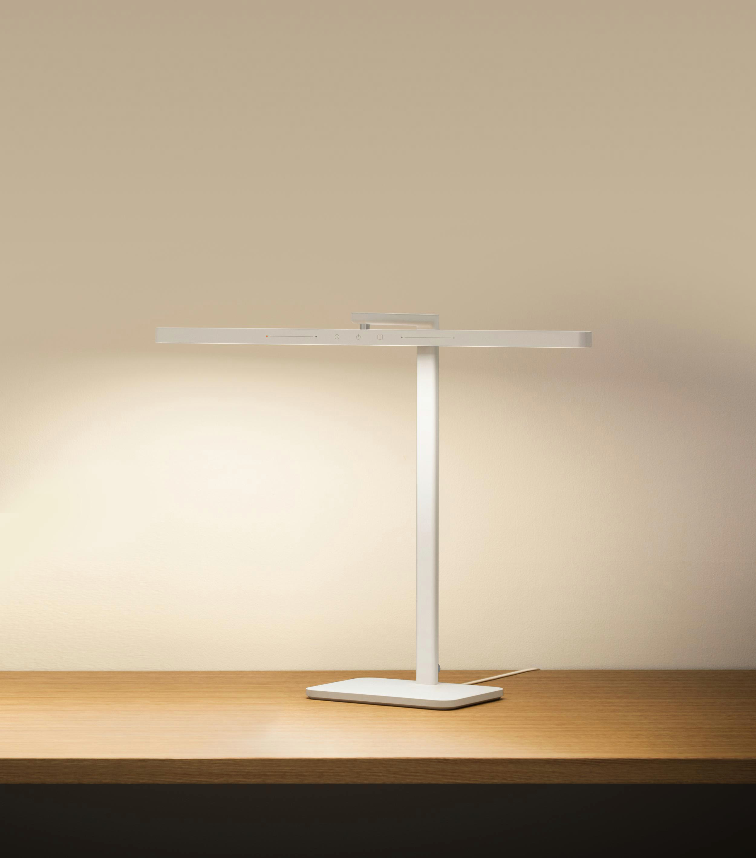 Eine weiße Schreibtischlampe auf einem Holztisch. Die Lampe hat ein langes, rechteckiges Licht und einen vertikalen Träger.