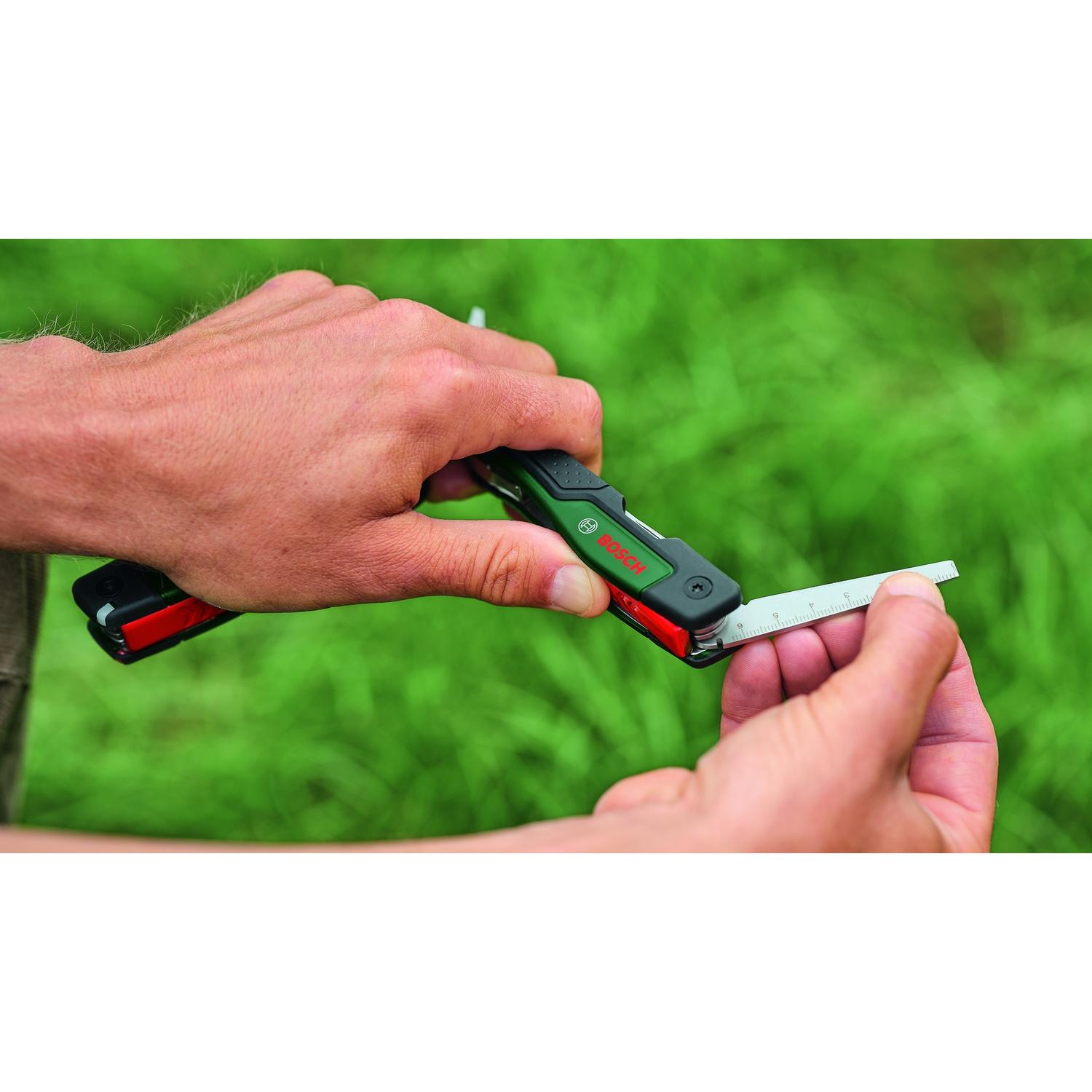 Mani che tengono un multiutensile verde e nero con un righello esteso. Erba verde sullo sfondo.