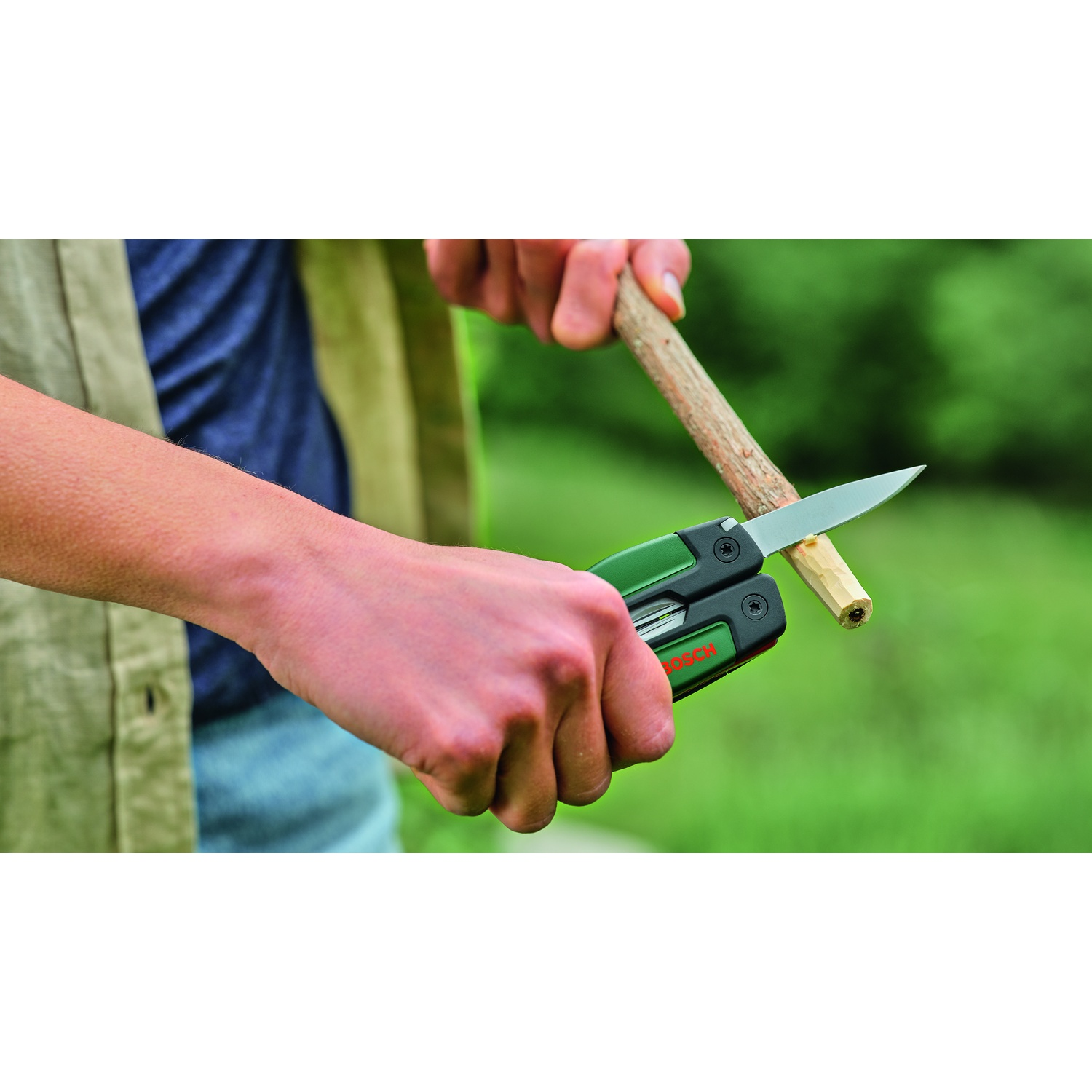 Una persona usa un coltello multiutensile verde e nero per intagliare un piccolo bastoncino di legno. Lo sfondo è un fogliame verde sfocato.