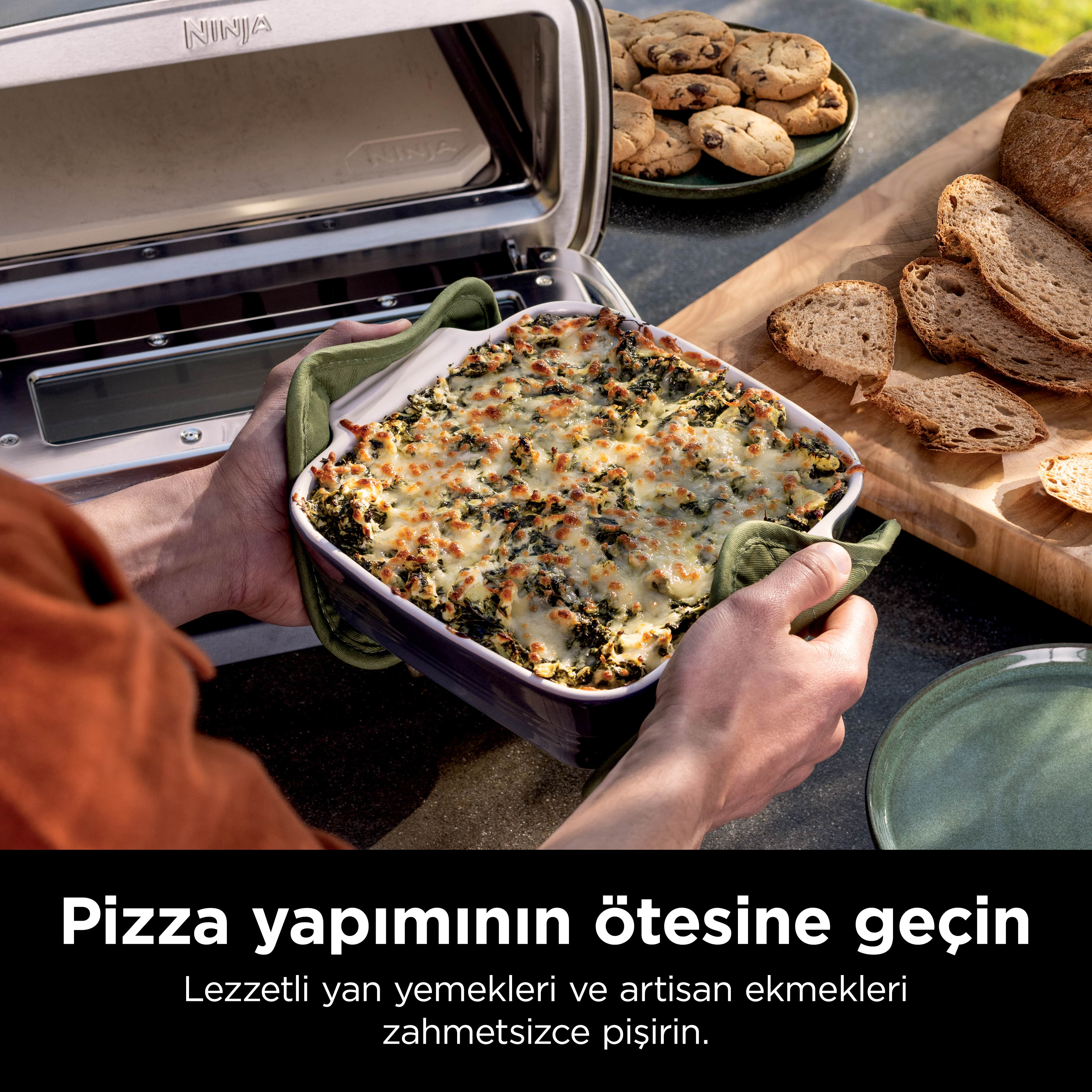 Bir kişi, eritilmiş peynirli fırınlanmış bir yemeği, ekmek, kurabiye ve bir Ninja fırınının yanında tutuyor. Yemeğe odaklan.