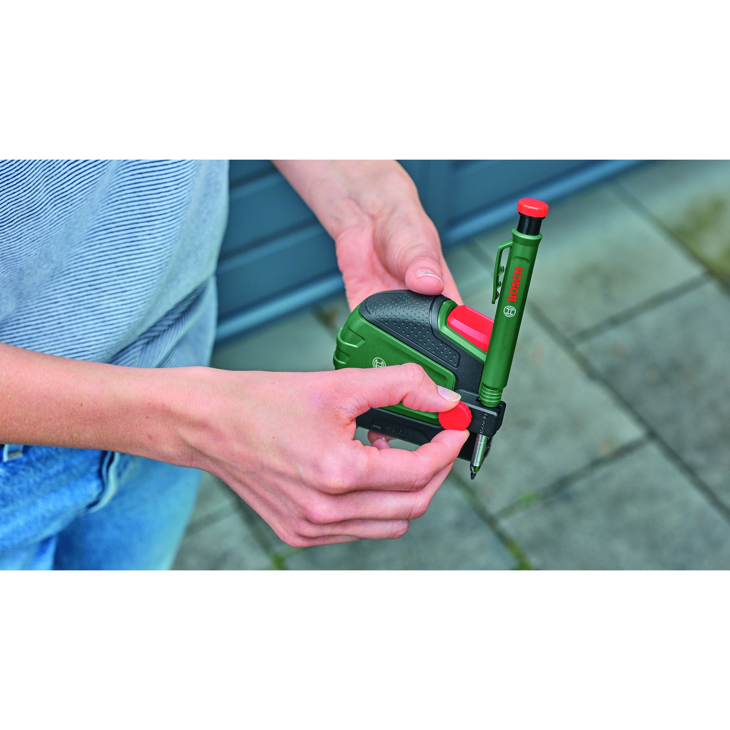 Una persona tiene in mano un utensile di marcatura Bosch verde e rosso. Le mani tengono l'utensile. Lo sfondo è una superficie piastrellata grigia.
