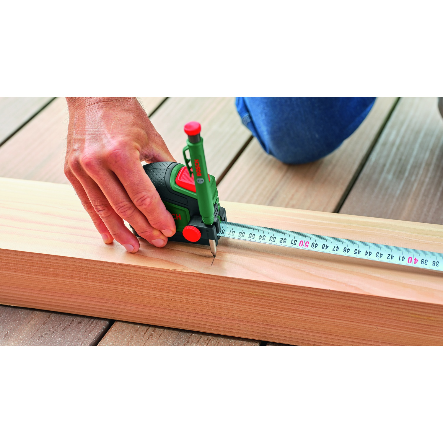 Una mano usa uno strumento di misura verde e nero per segnare un'asse di legno. Un metro a nastro si estende sul legno.
