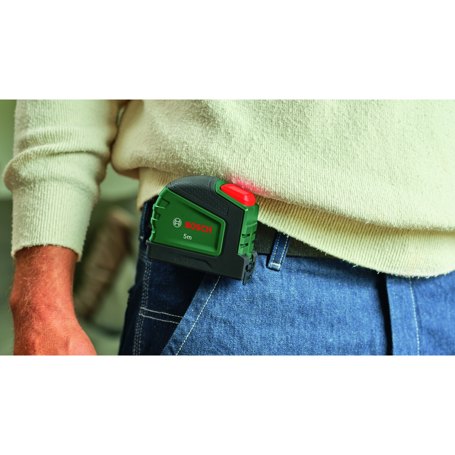 Una persona che indossa jeans e un maglione color crema ha un livella laser Bosch verde agganciata alla tasca. Il laser rosso è visibile.