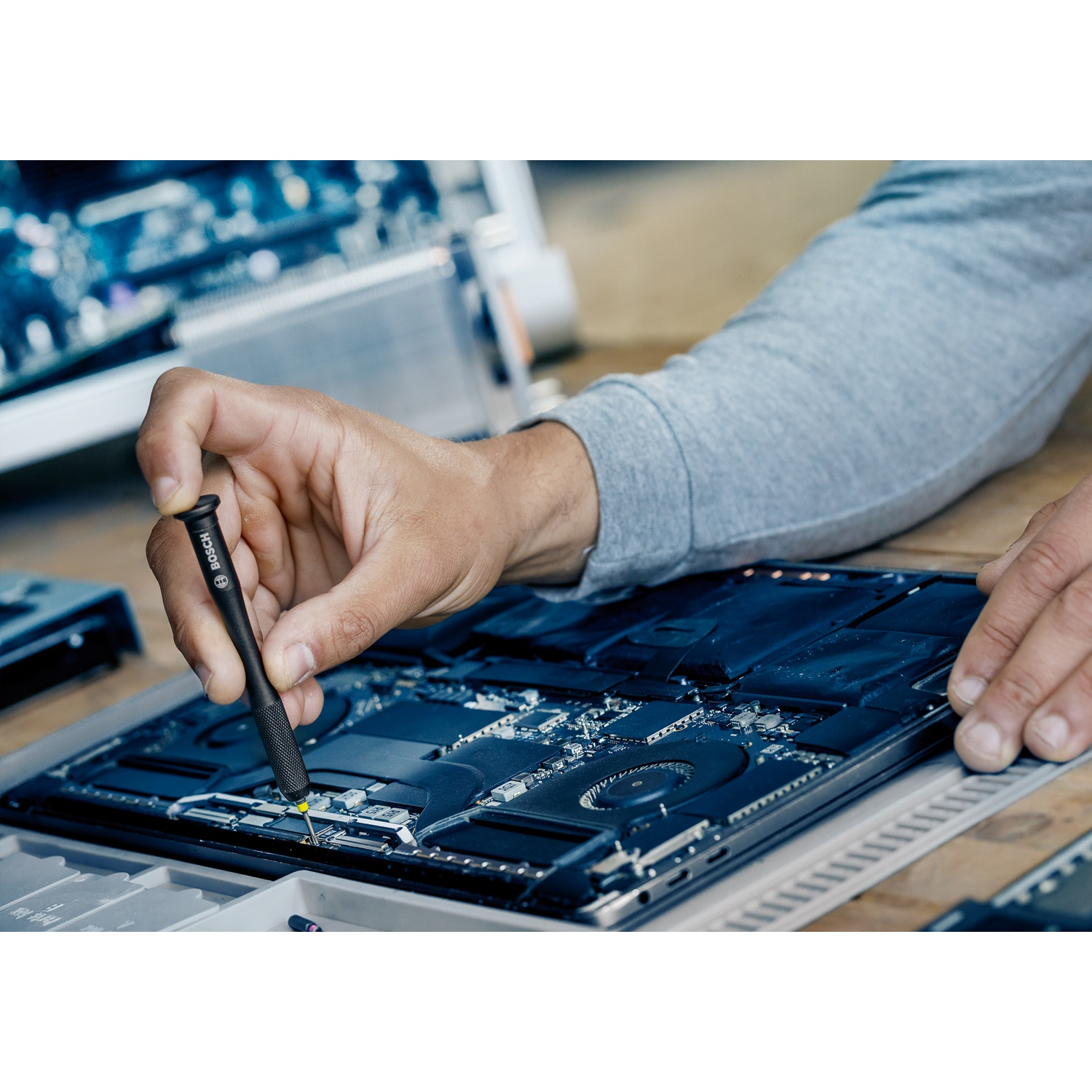 Una persona usa un cacciavite per riparare un laptop. I componenti interni del laptop sono visibili. Camicia grigia, tavolo di legno.