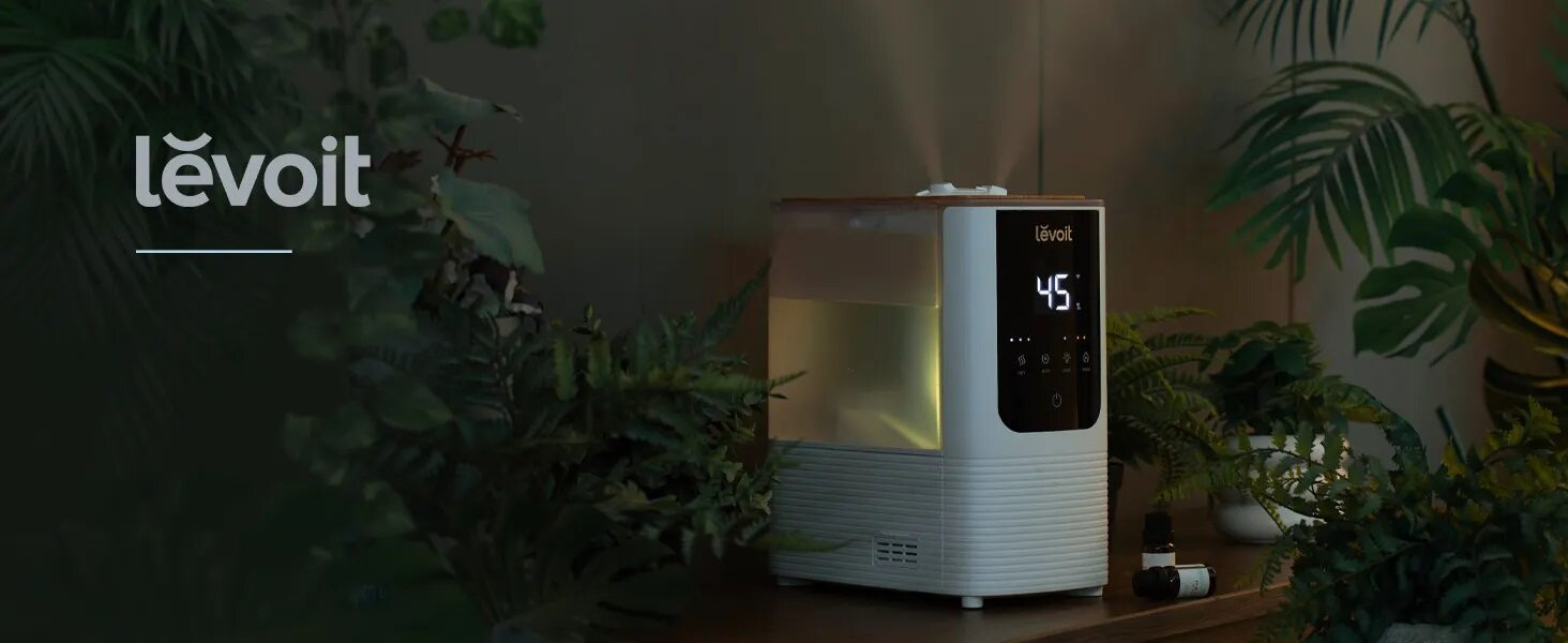 Nawilżacz powietrza Lëvoit z cyfrowym wyświetlaczem 45%. Otoczony zielonymi roślinami, na drewnianym stole mała butelka.