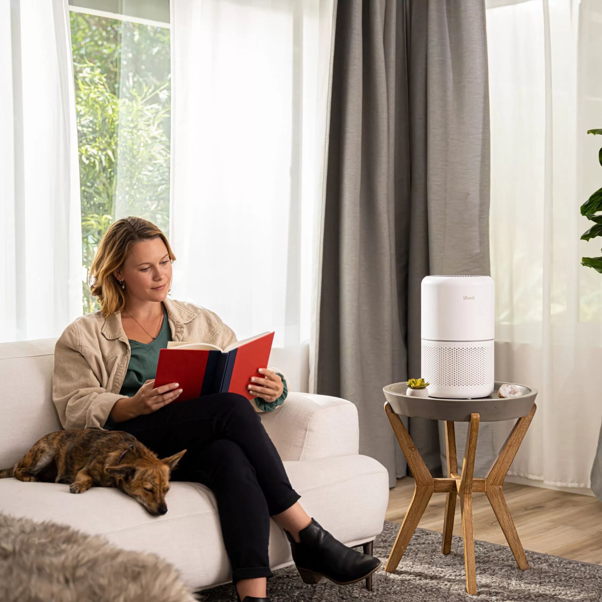 Kobieta czyta książkę na kanapie z psem. Biały oczyszczacz powietrza na stoliku. Zasłony i okno w tle.