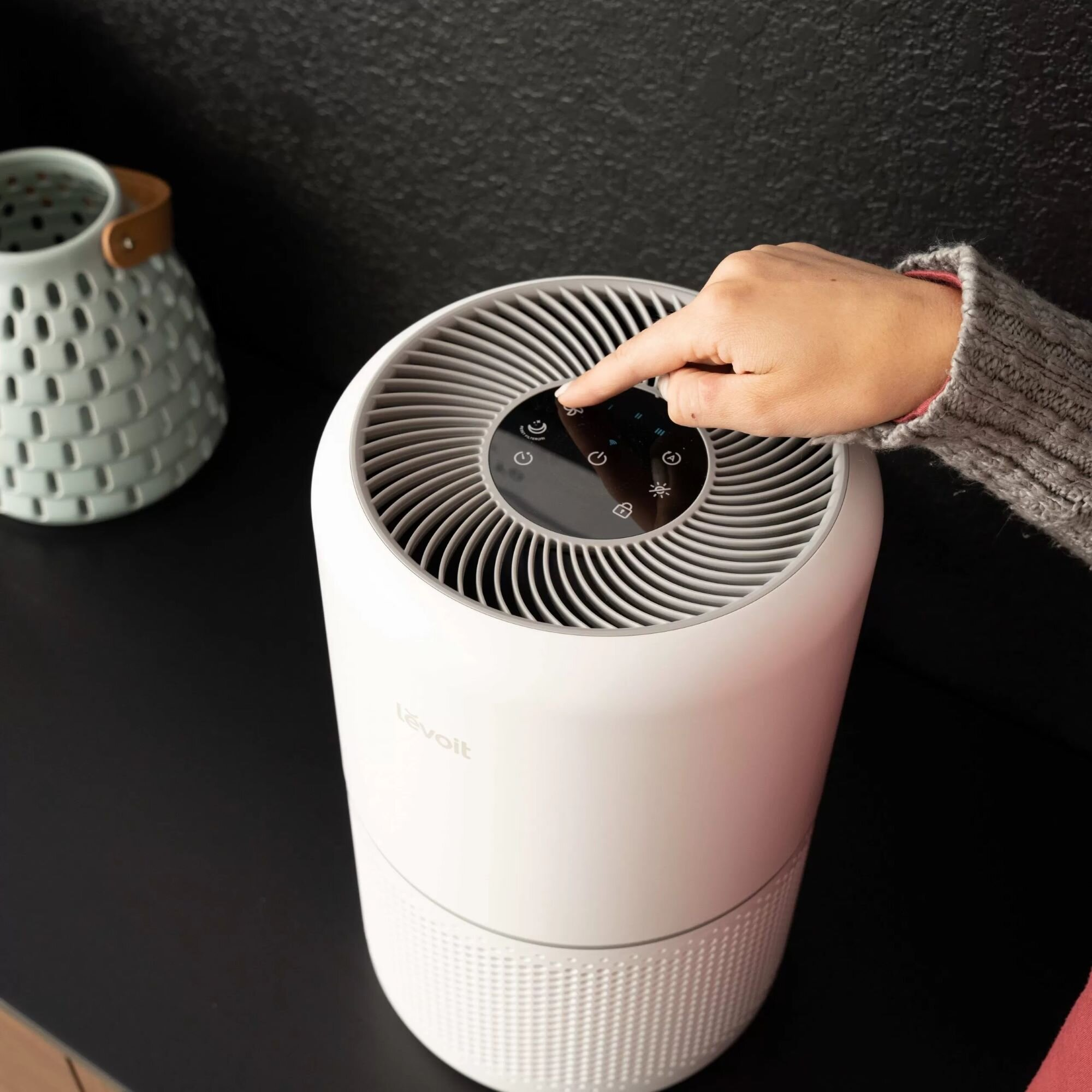 Ręka naciska przycisk na białym oczyszczaczu powietrza. Po lewej stronie znajduje się ozdobny wazon. Tło jest czarne.