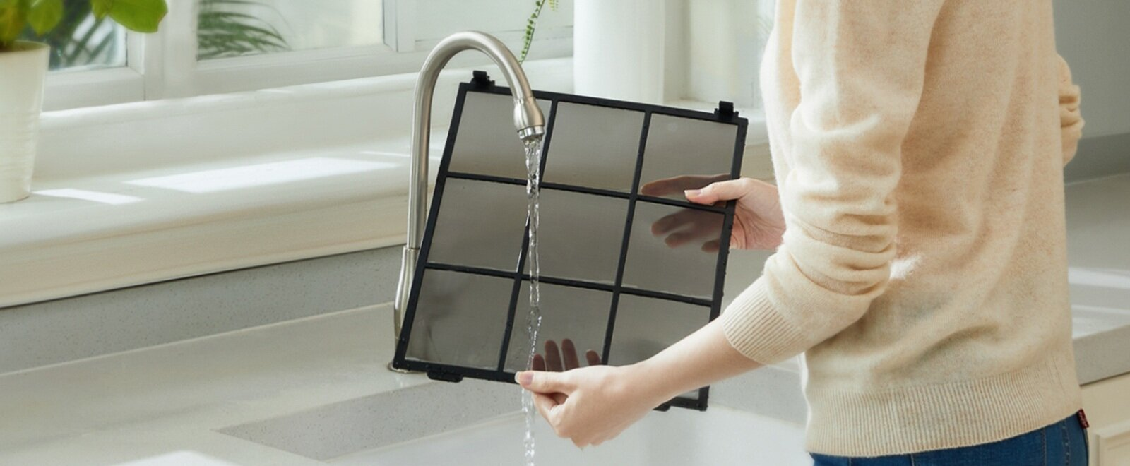 Osoba płucze filtr powietrza pod kranem. Filtr jest czarny, a osoba ma na sobie beżowy sweter i niebieskie dżinsy.