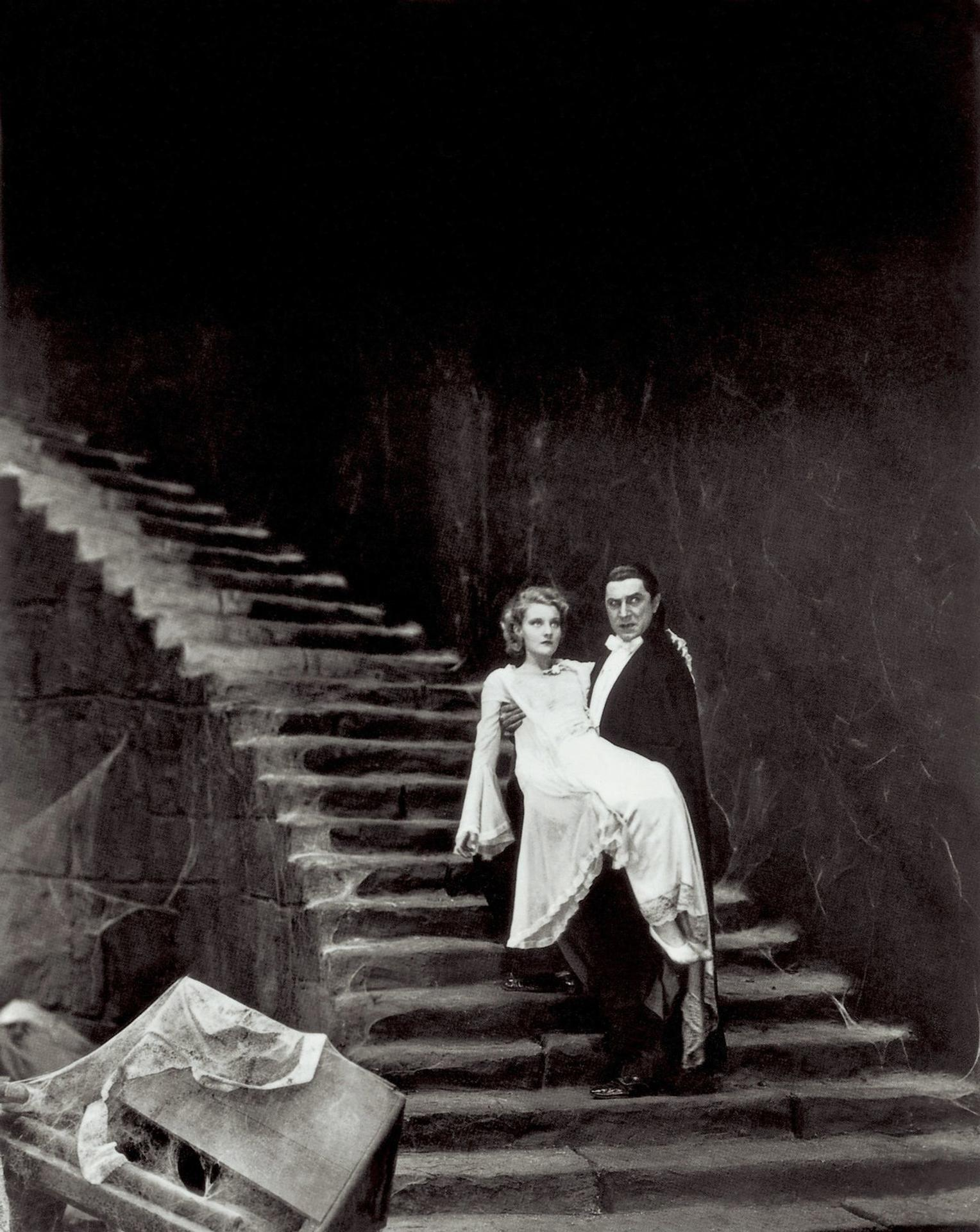 Ein Mann im Smoking trägt eine Frau in einem weißen Kleid eine Steintreppe hinunter. Ein Sarg steht in der Nähe, bedeckt mit Spinnweben.