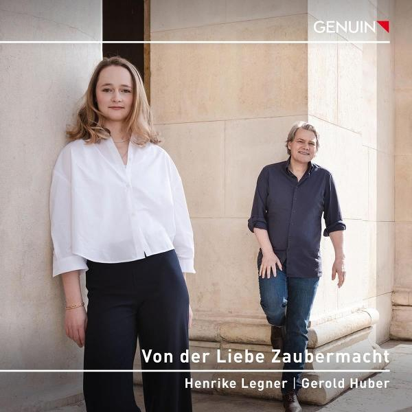Zwei Personen stehen an einem Gebäude. Eine Frau in weißem Hemd und schwarzer Hose und ein Mann in blauem Hemd und Jeans. Text: 'Von der Liebe Zaubermacht'.