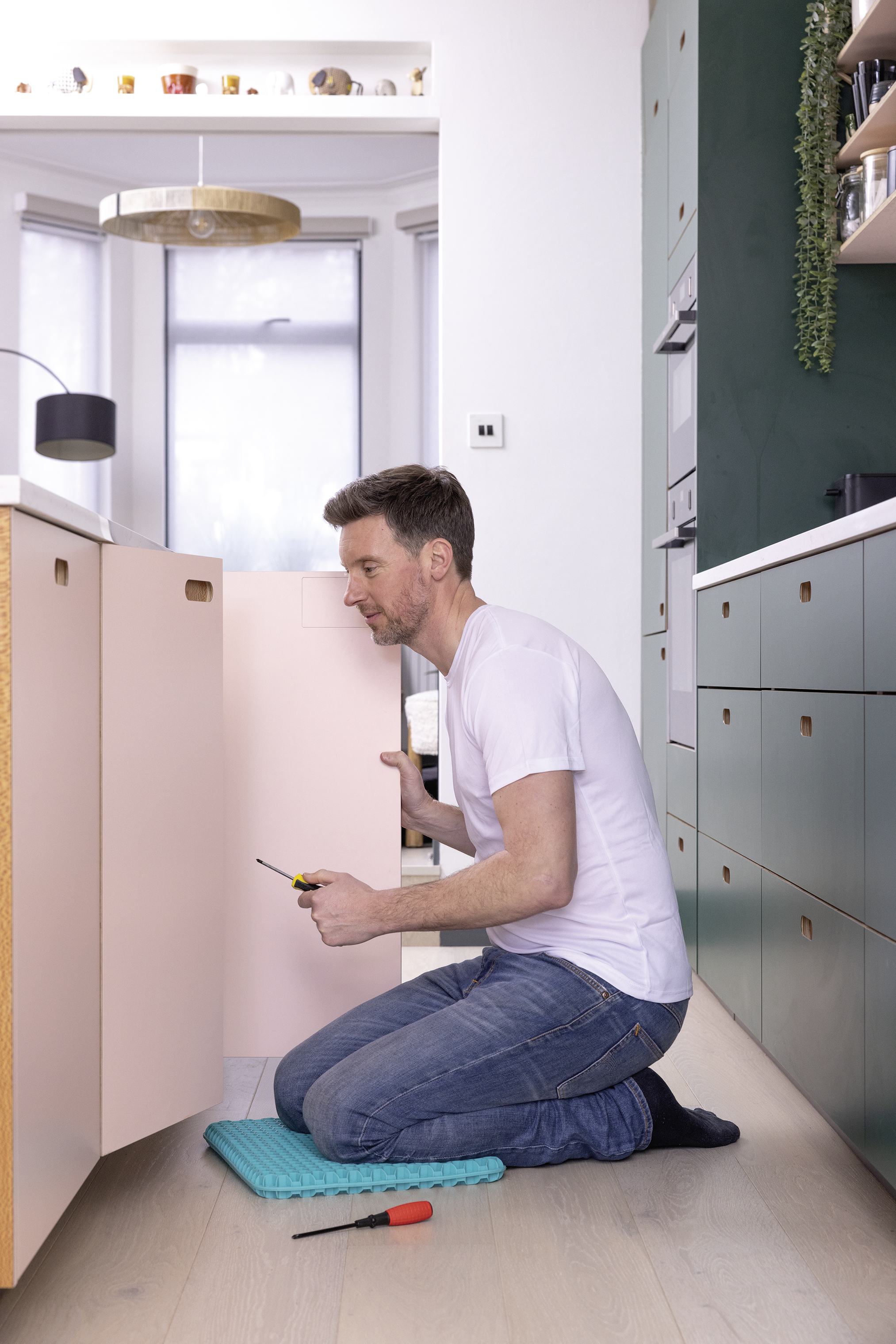 Mann kniet, schraubt an rosa Schrank. Blaues Kissen und roter Schraubenzieher daneben.
