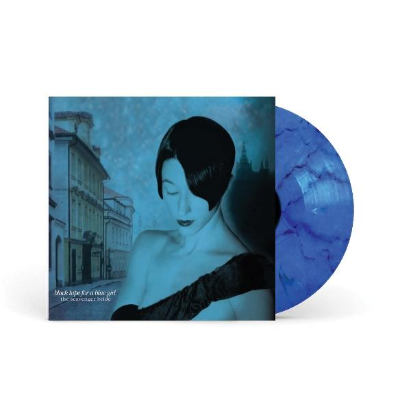 Albumcover mit einer Frau in Blau, schwarzen Haaren und Handschuhen. Eine blaue Schallplatte befindet sich rechts. Gebäude und Text sind sichtbar.