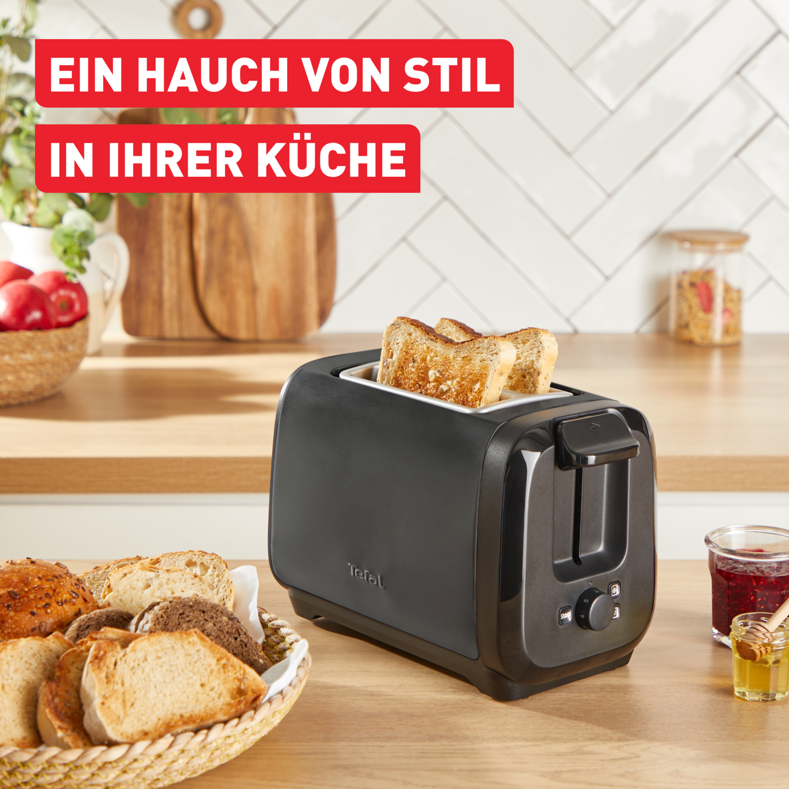 Ein schwarzer Toaster mit Toastbrotscheiben steht auf einer Holztheke. Ein Brotkorb, Marmelade und Honig sind ebenfalls vorhanden.