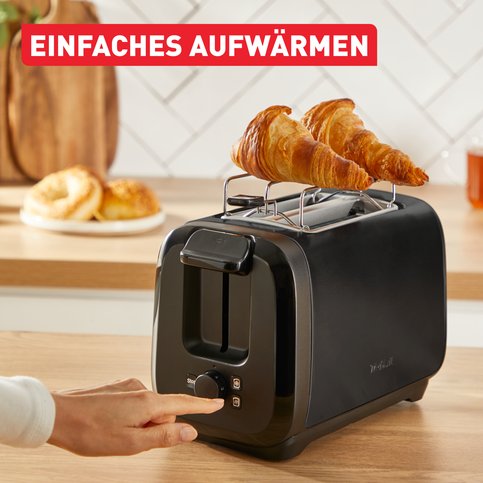 Ein schwarzer Toaster mit zwei Croissants. Eine Hand drückt eine Taste. Bagels sind im Hintergrund.