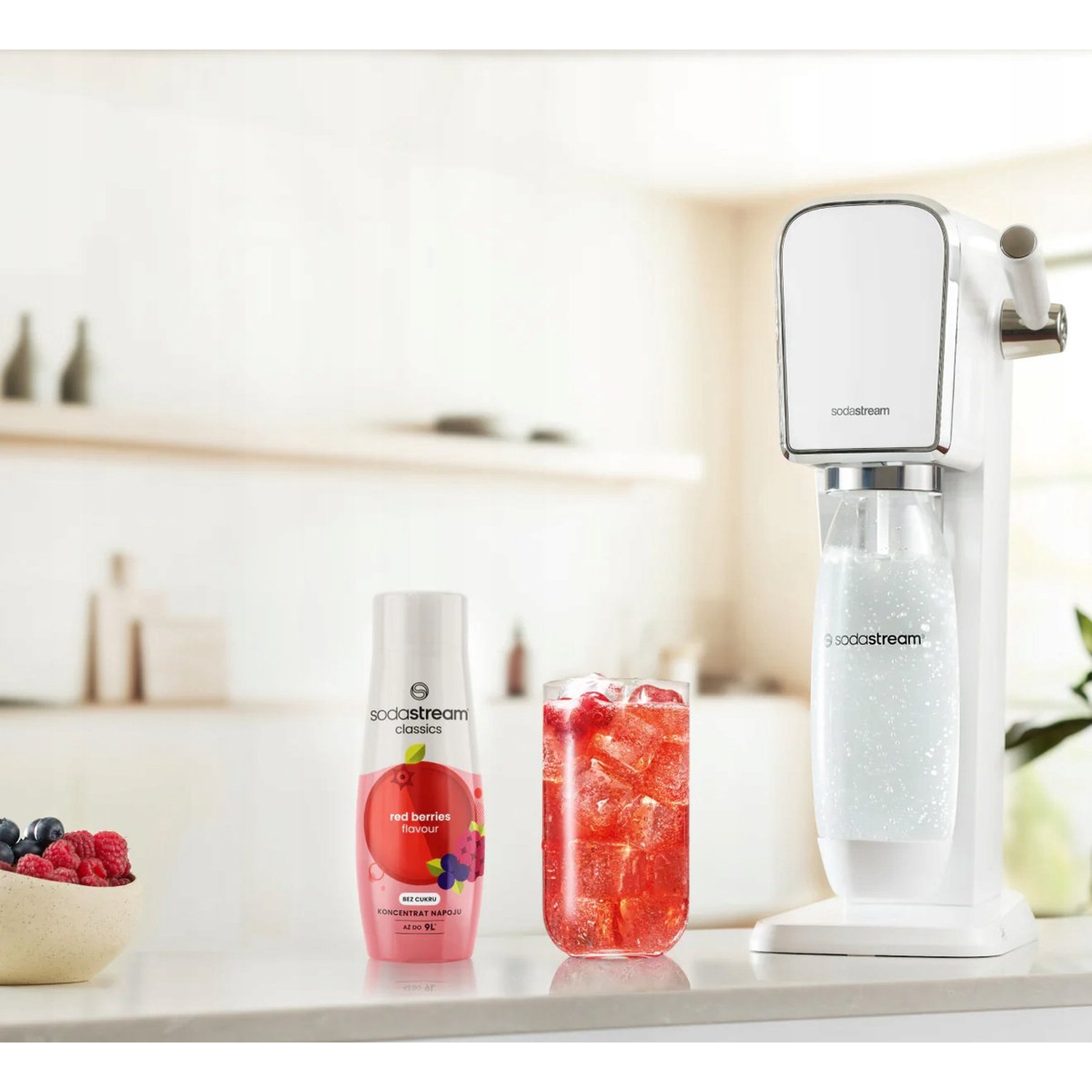 A SodaStream machine, a red berry drink, a red berries syrup, and a bowl of berries are on a white surface.