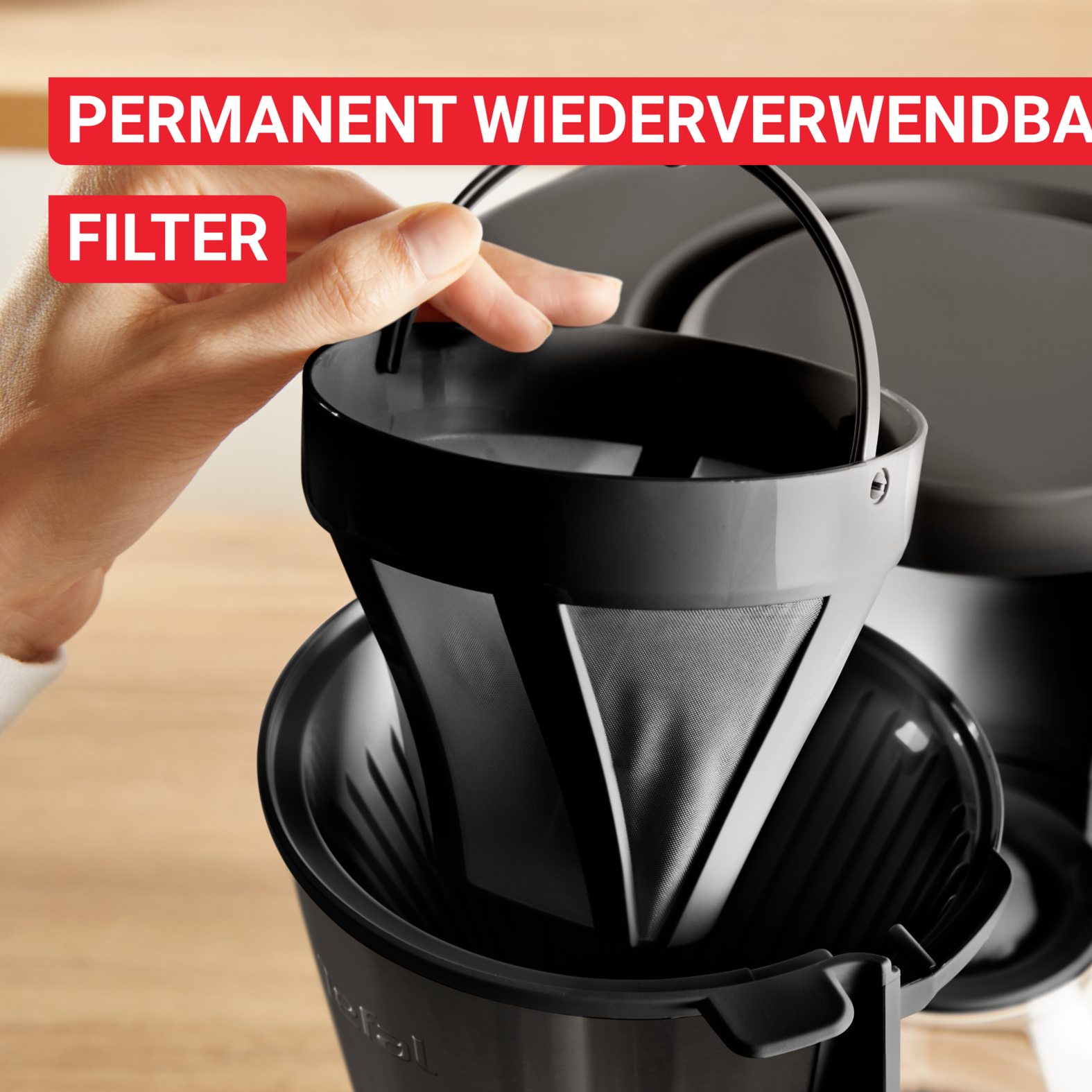 Eine Hand hält einen schwarzen Kaffeefilter über einer Kaffeemaschine. Die Wörter 'PERMANENT WIEDERVERWENDBAR' und 'FILTER' sind sichtbar.