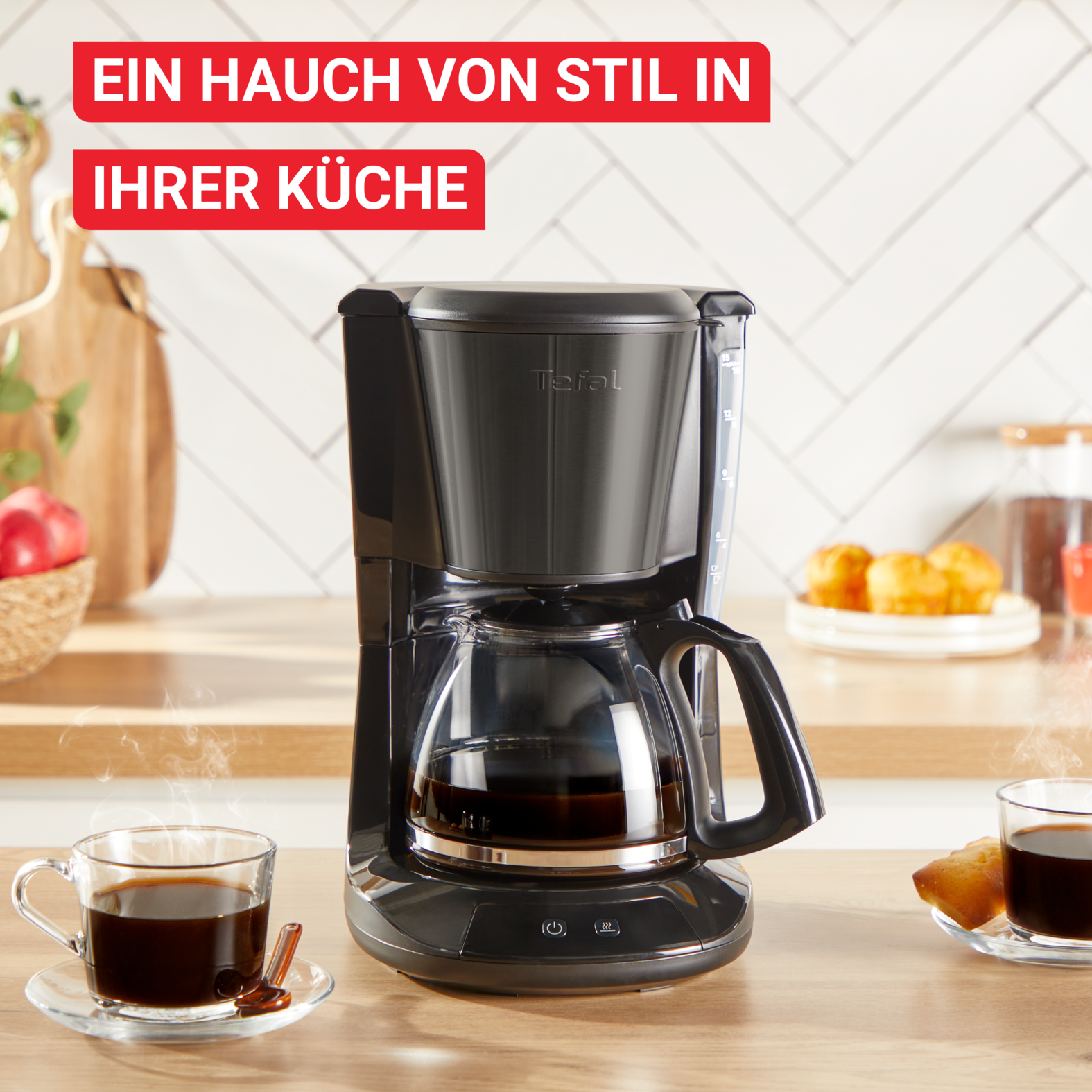 Eine schwarze Kaffeemaschine mit Glaskaraffe, zwei Kaffeetassen und Gebäck auf einem Holztisch. Text oben: Ein Hauch von Stil.