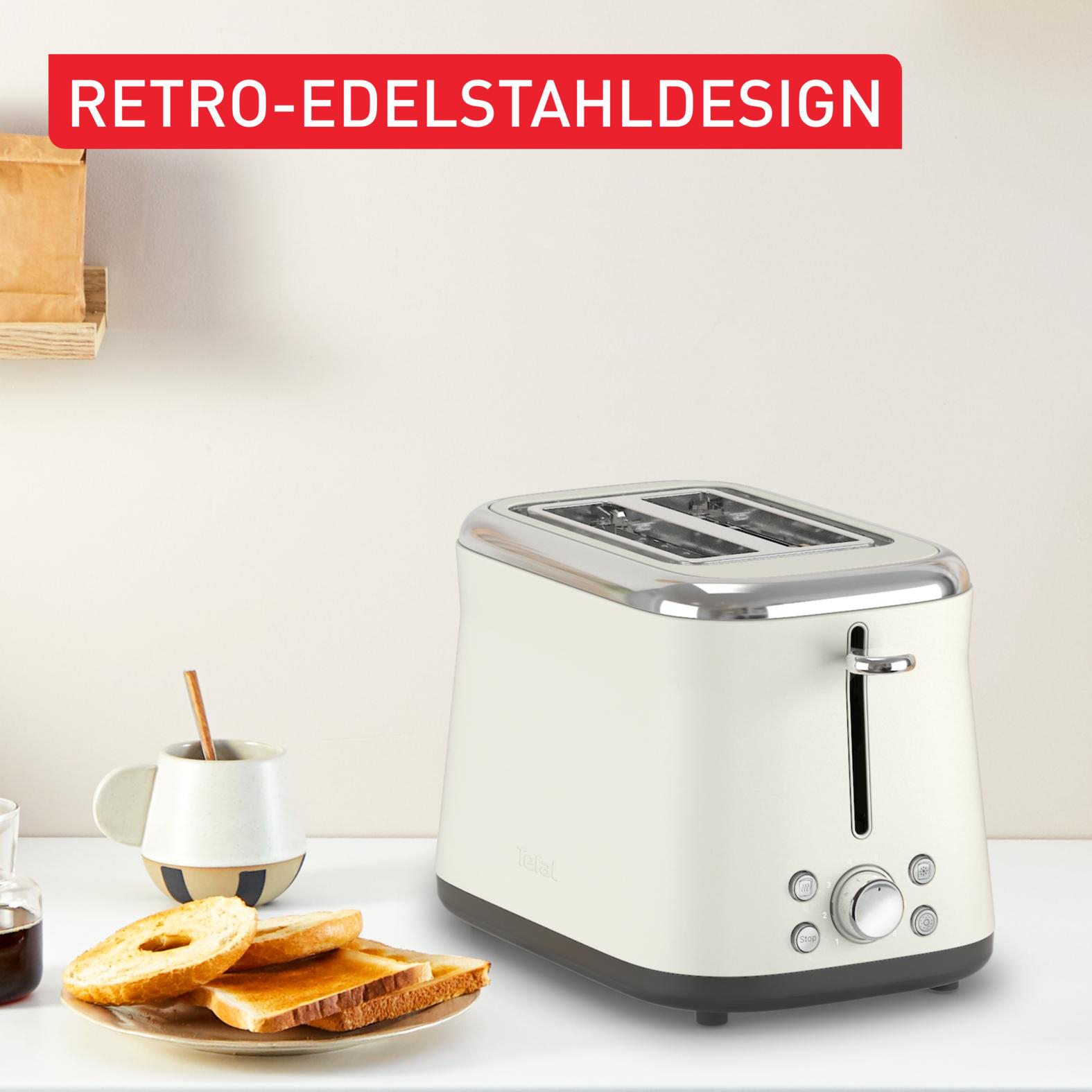 Ein cremefarbener Retro-Toaster mit Akzenten aus Edelstahl steht auf einer weißen Oberfläche neben Toast und einer Tasse.