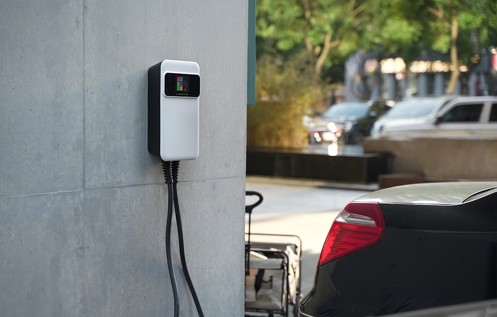 Egy elektromos autótöltő egy betonfalon. Egy fekete autó parkol a közelben. Fák és más autók a háttérben.