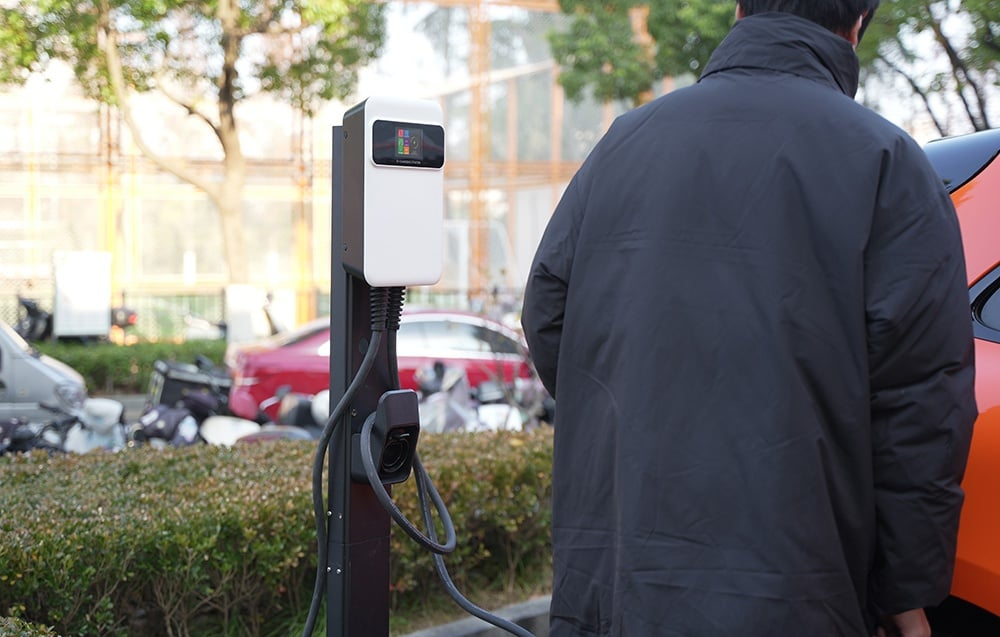 Egy személy egy elektromos jármű töltőállomásánál áll. Autók és robogók a homályos háttérben. Egy narancssárga autó látható.