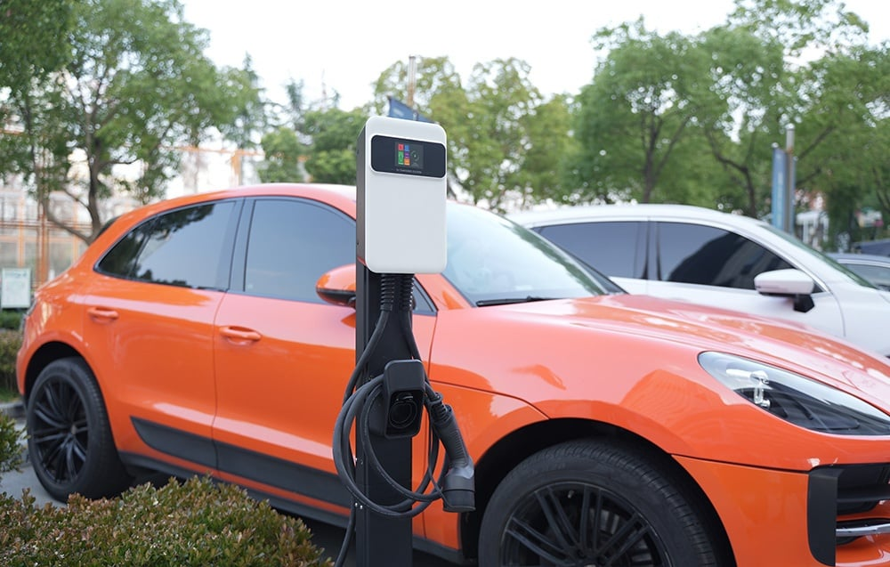 Egy narancssárga autó töltődik egy elektromos autó töltőállomásán. Mögötte egy fehér autó parkol. Zöld fák a háttérben.