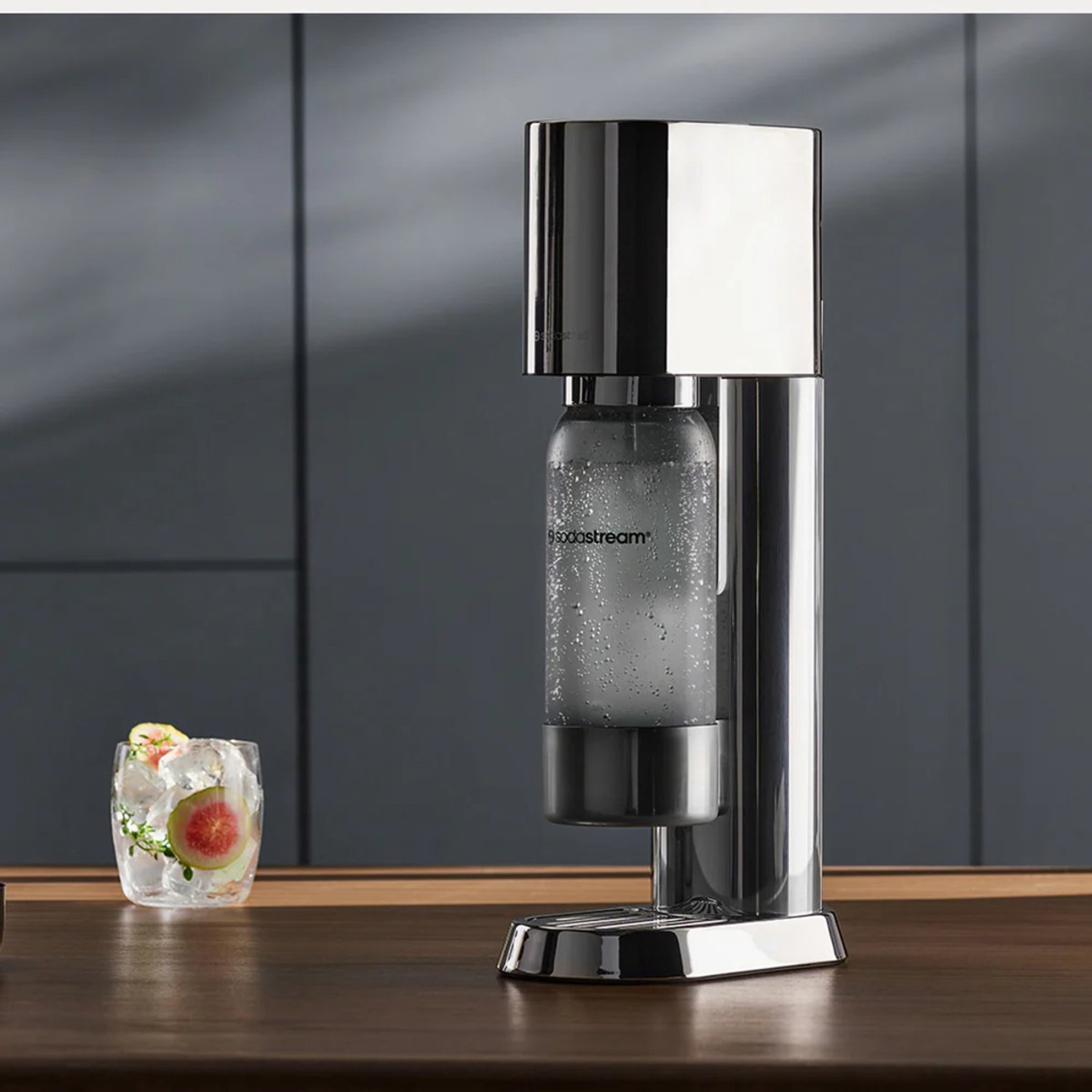 A chrome SodaStream machine with a glass of cocktail on a wooden table. Dark grey background.