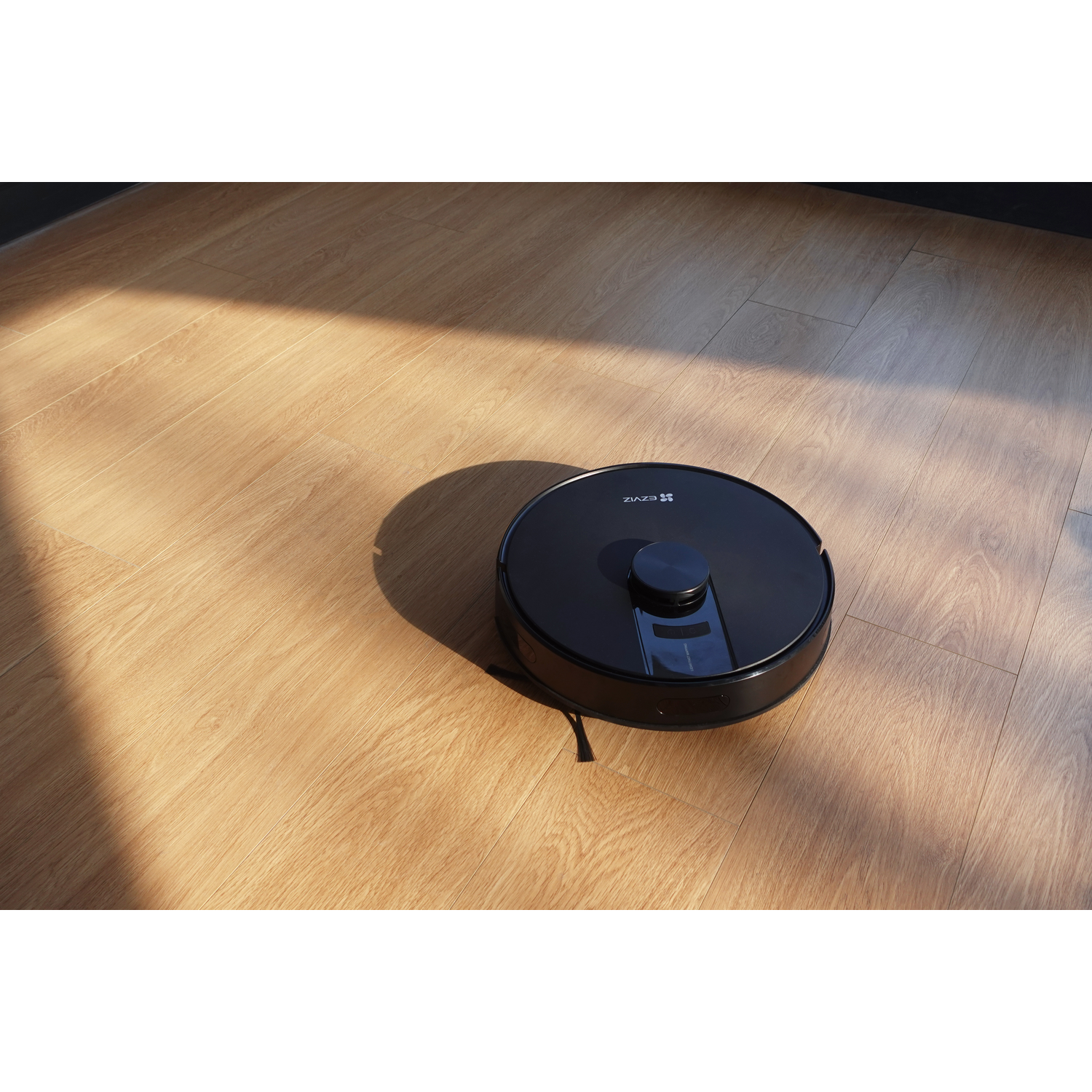 Een zwarte robotstofzuiger op een houten vloer, gedeeltelijk verlicht door zonlicht. De stofzuiger heeft een ronde vorm en de merknaam is zichtbaar.