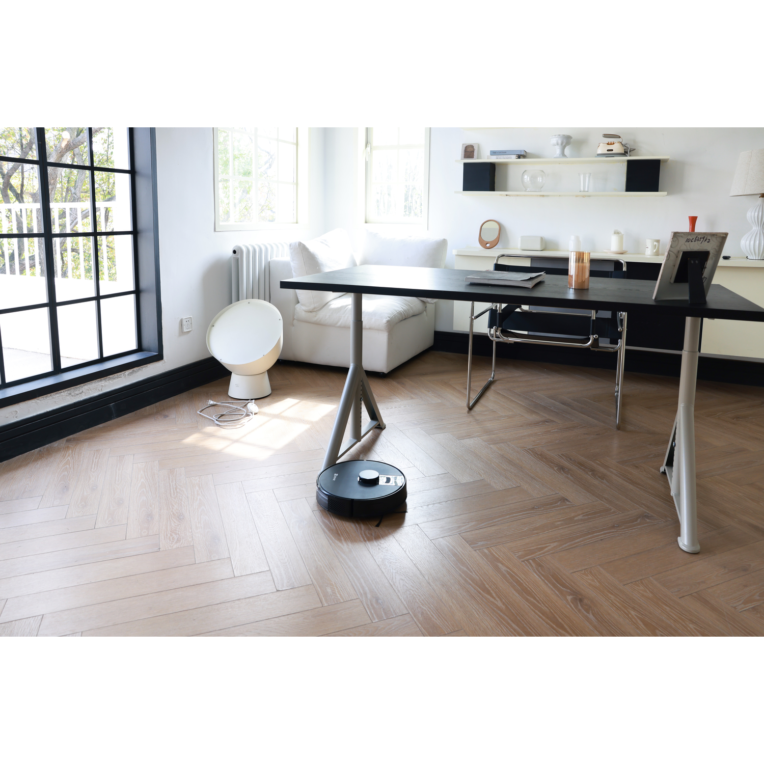 Een zwarte robotstofzuiger maakt een kamer met een houten vloer schoon. Een bureau, bank en raam staan op de achtergrond.